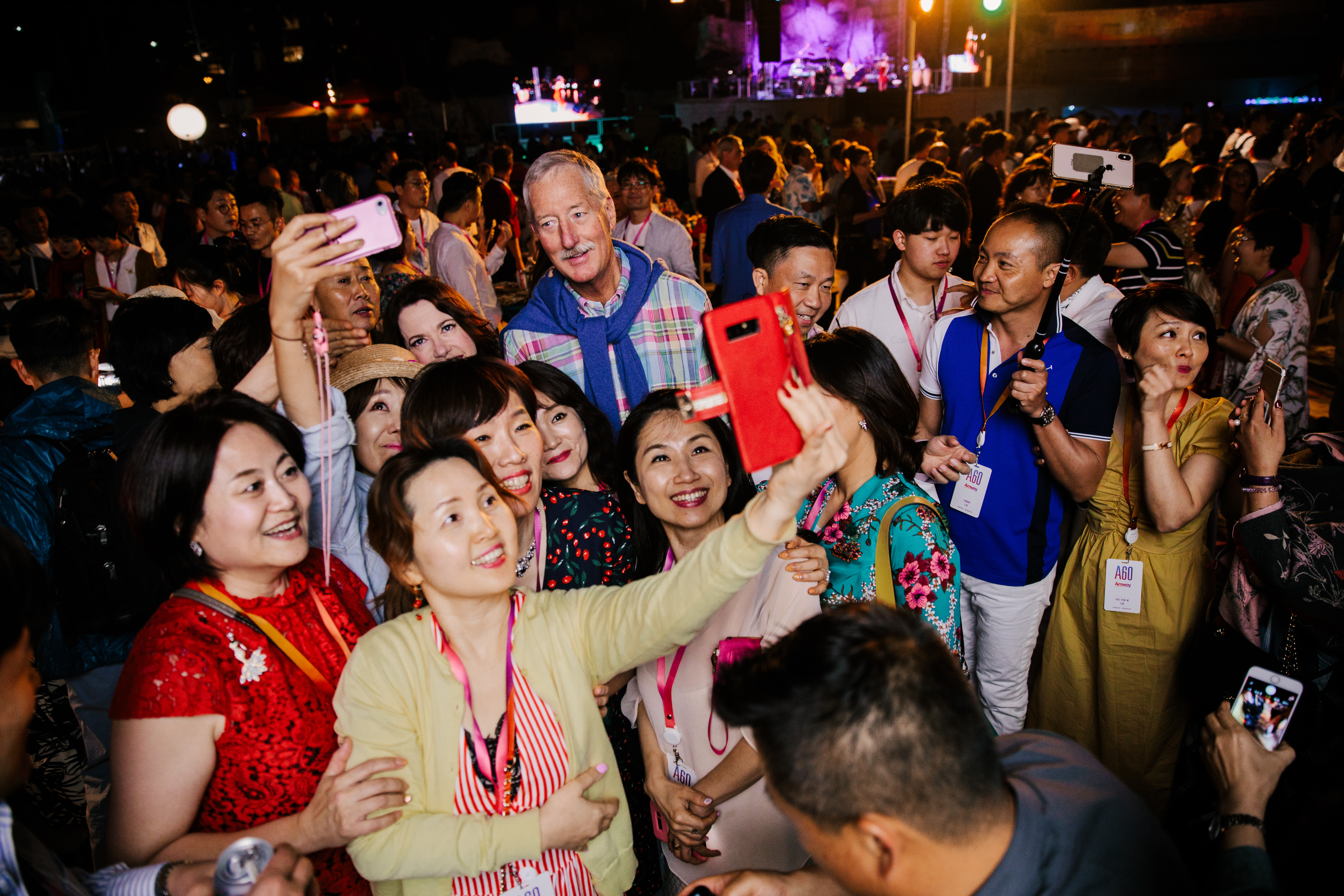 活动现场氛围热烈愉悦，参与人员均佩戴印有“AGO”字样的身份胸牌，大家簇拥着外籍来宾，纷纷举起手机自拍留念，人人脸上都洋溢着灿烂的笑容，背景舞台灯光璀璨，全场充满融洽欢乐的氛围，尽显安利伙伴的活力与团队凝聚力。