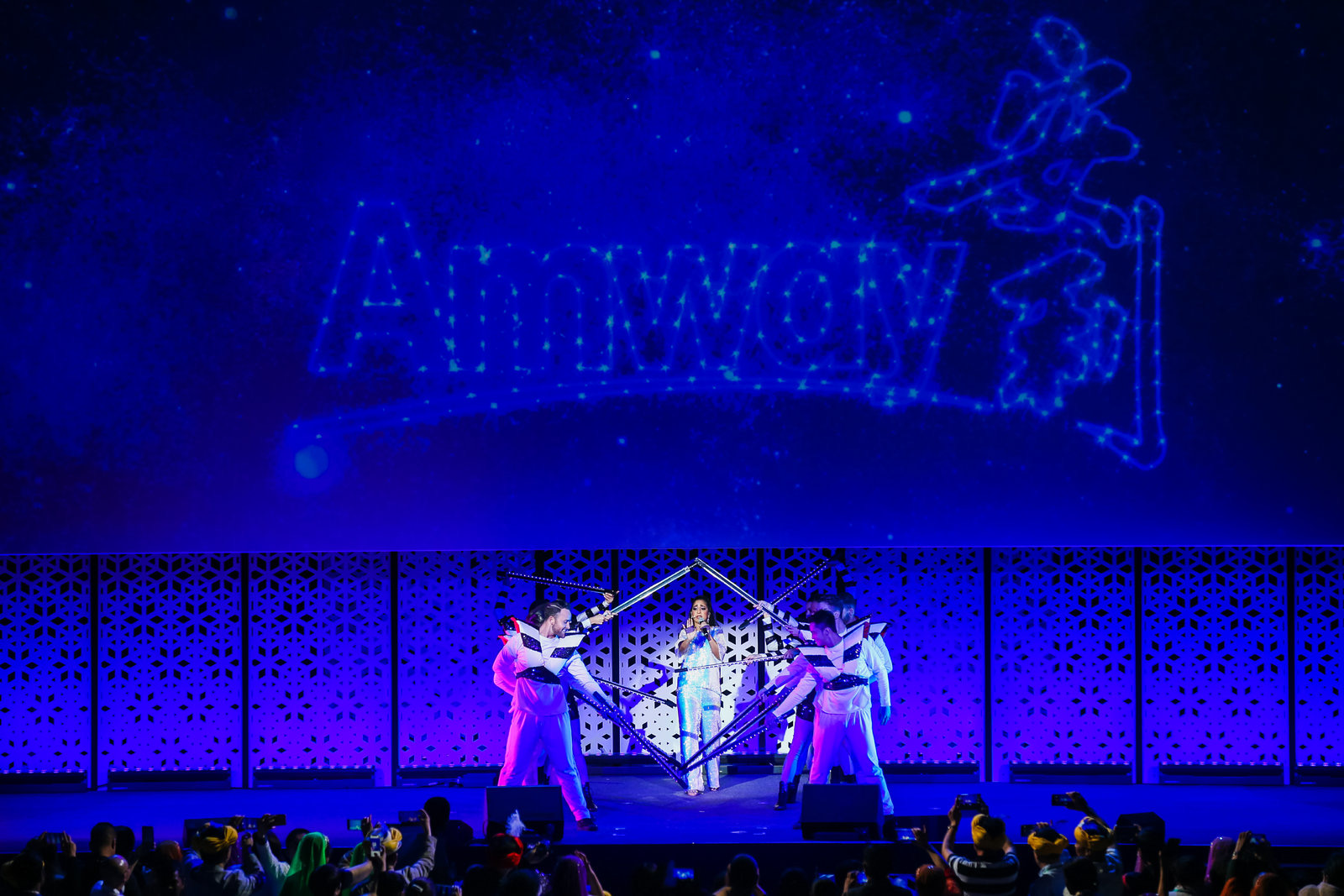 背景大屏呈现星光效果的“Amway 安利”字样，蓝紫色灯光氛围感拉满。舞台中央女歌手正倾情演唱，多名舞者持创意道具配合表演，编排精巧。台下观众纷纷举起手机记录精彩时刻，现场氛围热烈鲜活，尽显安利活动的活力质感。