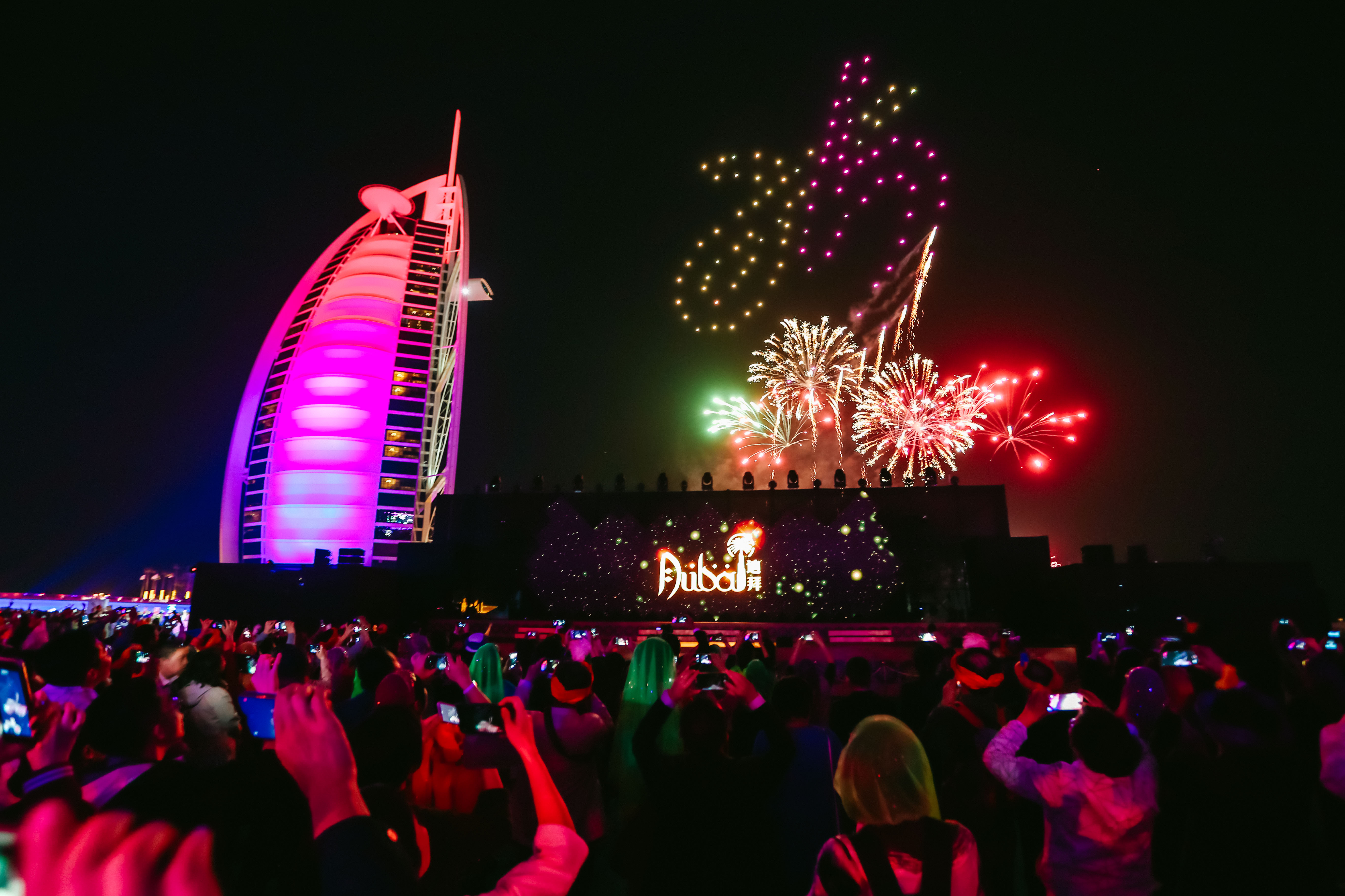 本次安利迪拜活动现场，大屏显示“Dubai迪拜”字样，旁侧被粉紫色灯光照亮的帆船酒店醒目亮眼，夜空中多彩烟花次第绚烂绽放，台下参会的安利伙伴纷纷举起手机记录盛景，现场氛围热烈欢腾，尽显本次活动的盛大与荣耀。