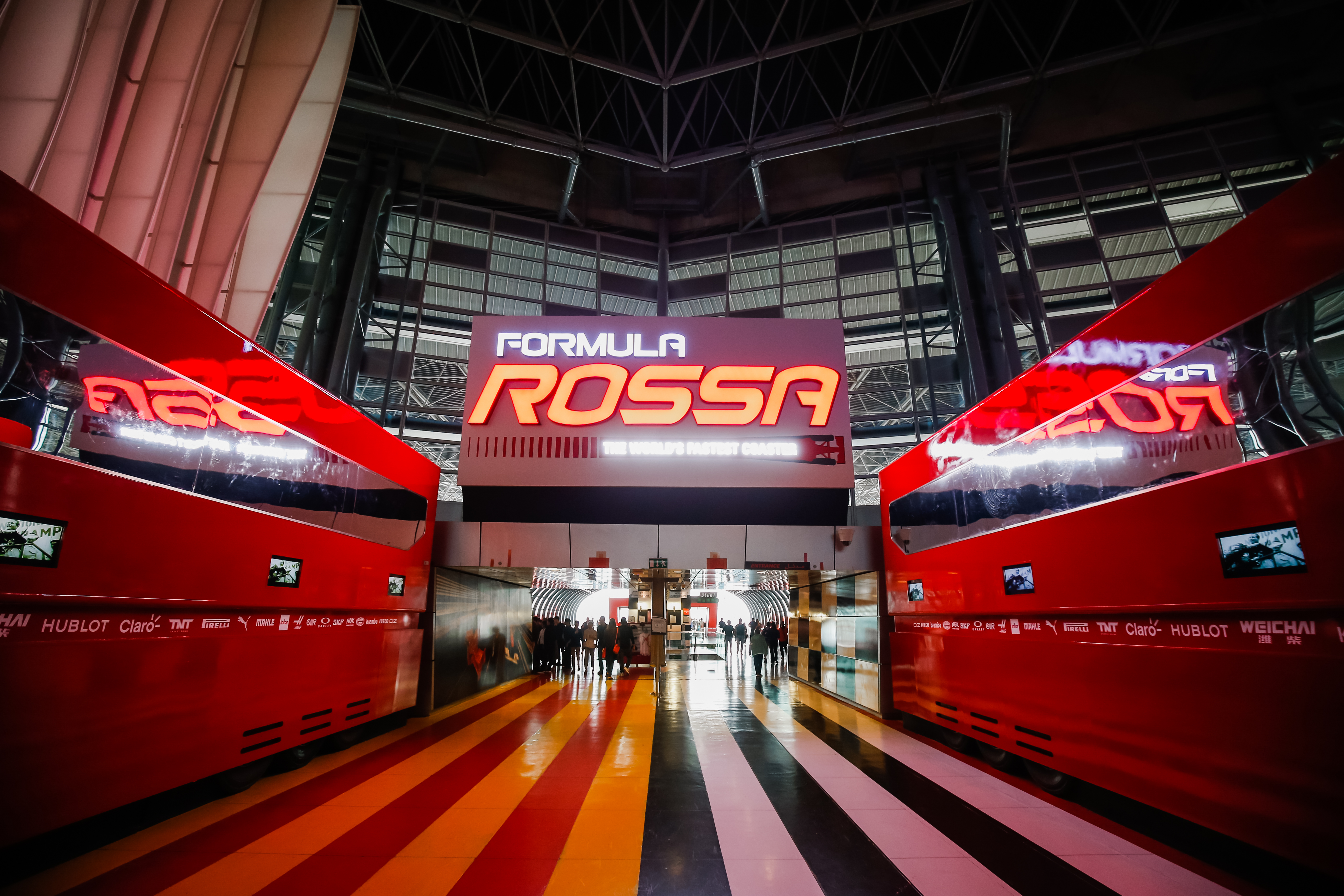 画面核心标识为“FORMULA ROSSA”，标注有“THE WORLD'S FASTEST COASTER”字样，两侧红色围挡搭配撞色条纹地面，参会人员正有序步入通道，整体氛围动感热血，充满速度感与活力，契合品牌积极向上的宣传调性。