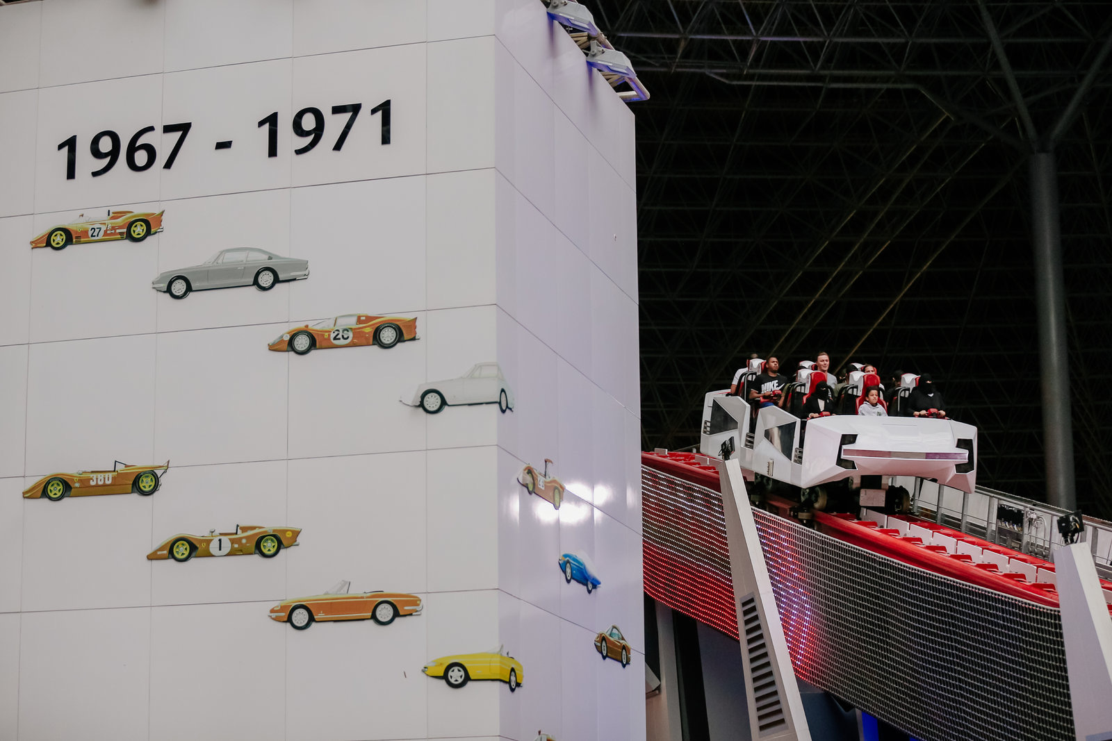 画面左侧立面标注核心文字“1967-1971”，搭配多款复古赛车造型装饰对应品牌发展阶段，右侧多位安利伙伴乘坐互动体验设施，氛围轻松鲜活，尽显品牌向上发展的活力与朝气。