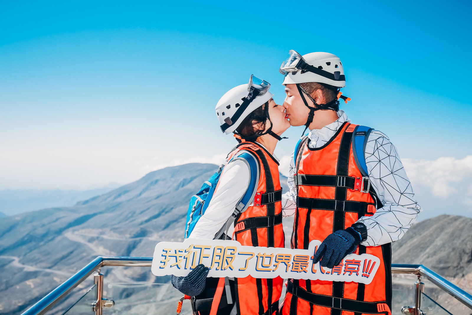 晴日蓝天之下，身着全套滑索安全护具的两位安利伙伴站在山巅观景台，手持写有“我征服了世界最长滑索！”的手举牌亲密亲吻留念，背后是壮阔连绵的群山，氛围甜蜜昂扬，尽显挑战自我的喜悦与活动的鲜活能量。