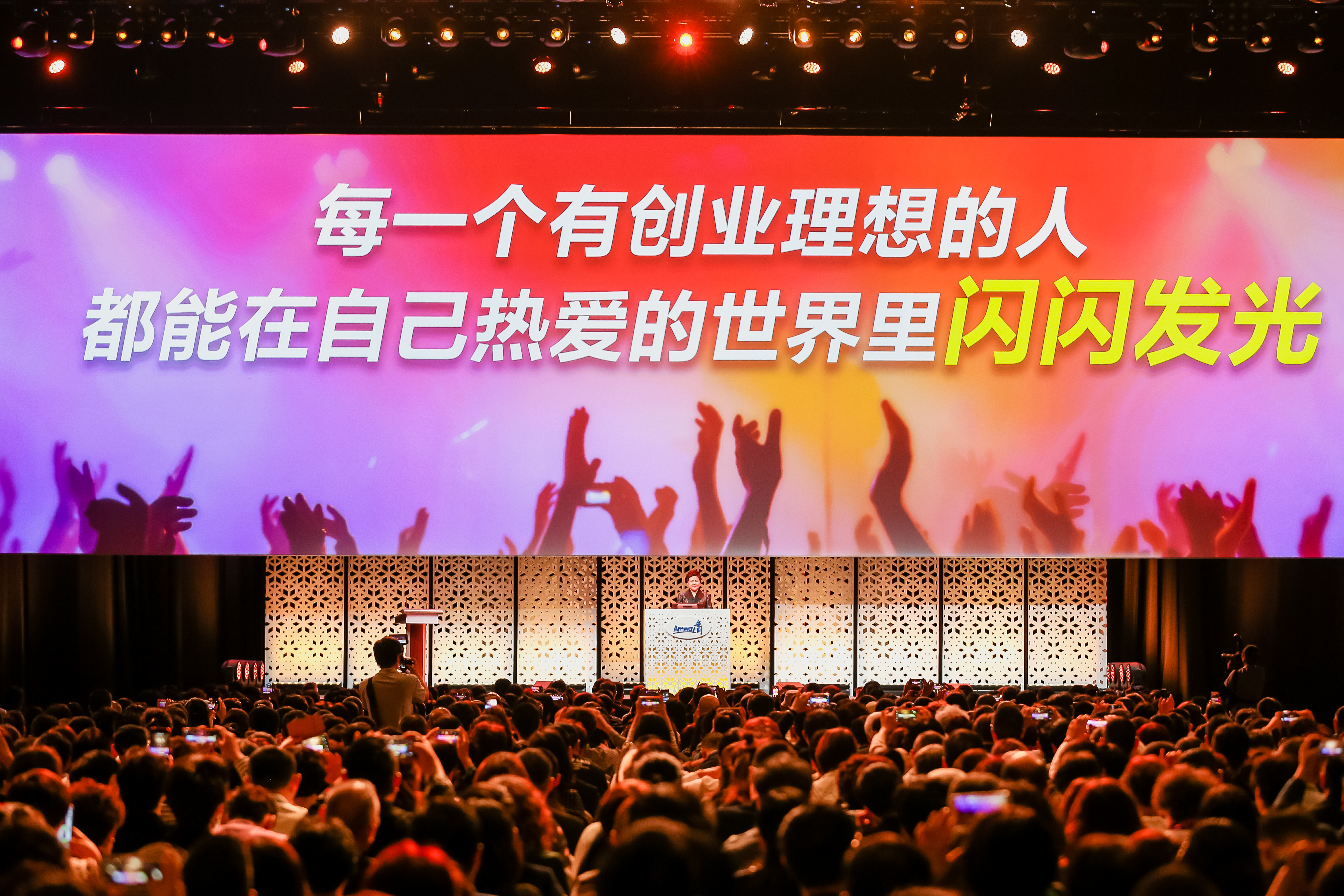 活动现场的巨型屏幕展示着标语“每一个有创业理想的人，都能在自己热爱的世界里闪闪发光”，嘉宾站在印有安利标识的讲台分享，台下坐满参会伙伴，大家纷纷举起手机记录内容，全场氛围热烈昂扬，传递出安利鼓励创业、支持伙伴逐梦的品牌理念。