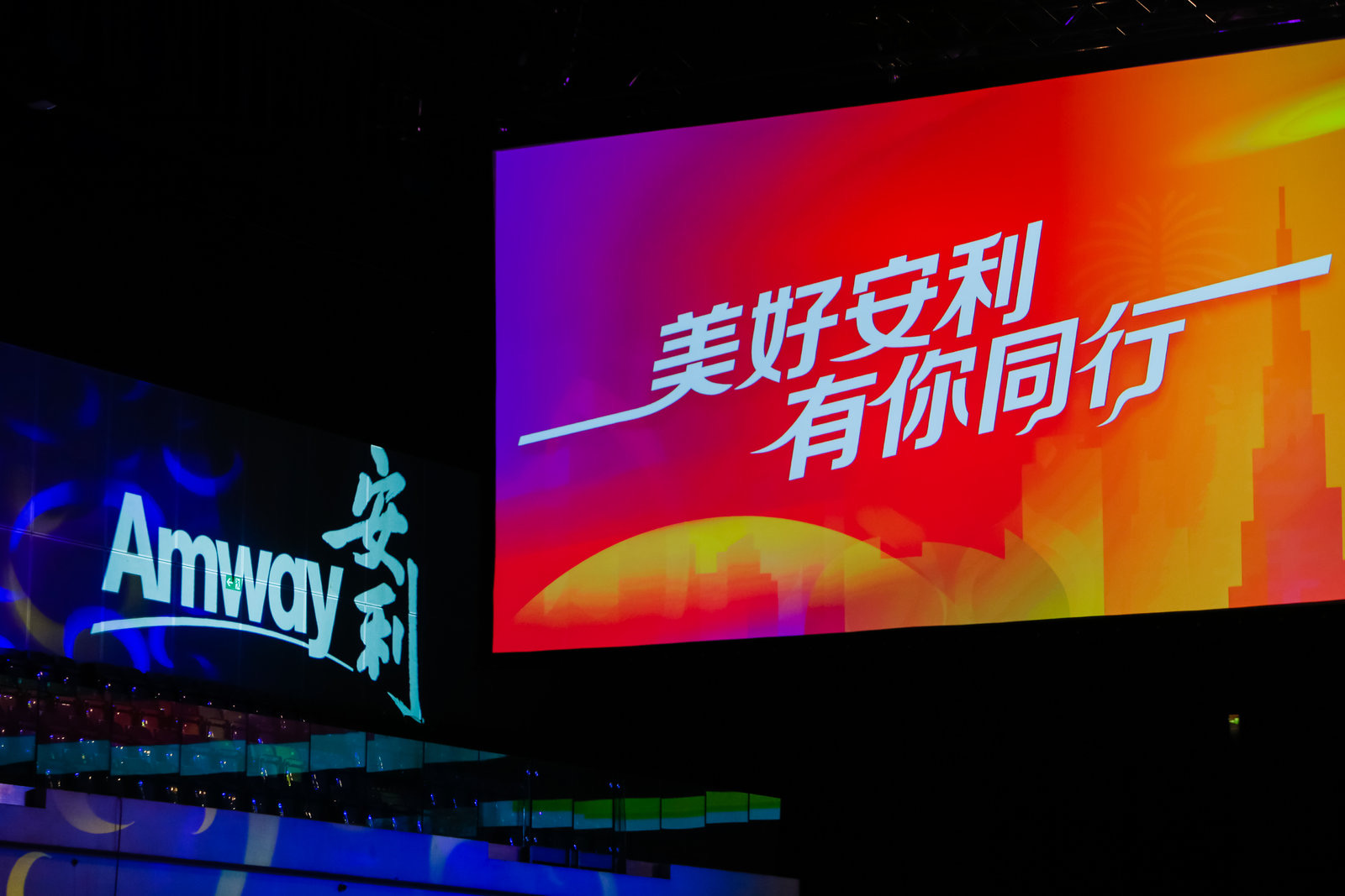 活动现场设置两块宣传大屏，左侧大屏为蓝紫色调，清晰呈现“Amway”及“安利”品牌标识；右侧大屏采用橙红渐变背景，醒目展示“美好安利 有你同行”主题标语，整体氛围隆重热烈，传递出温暖正向的品牌活动调性。