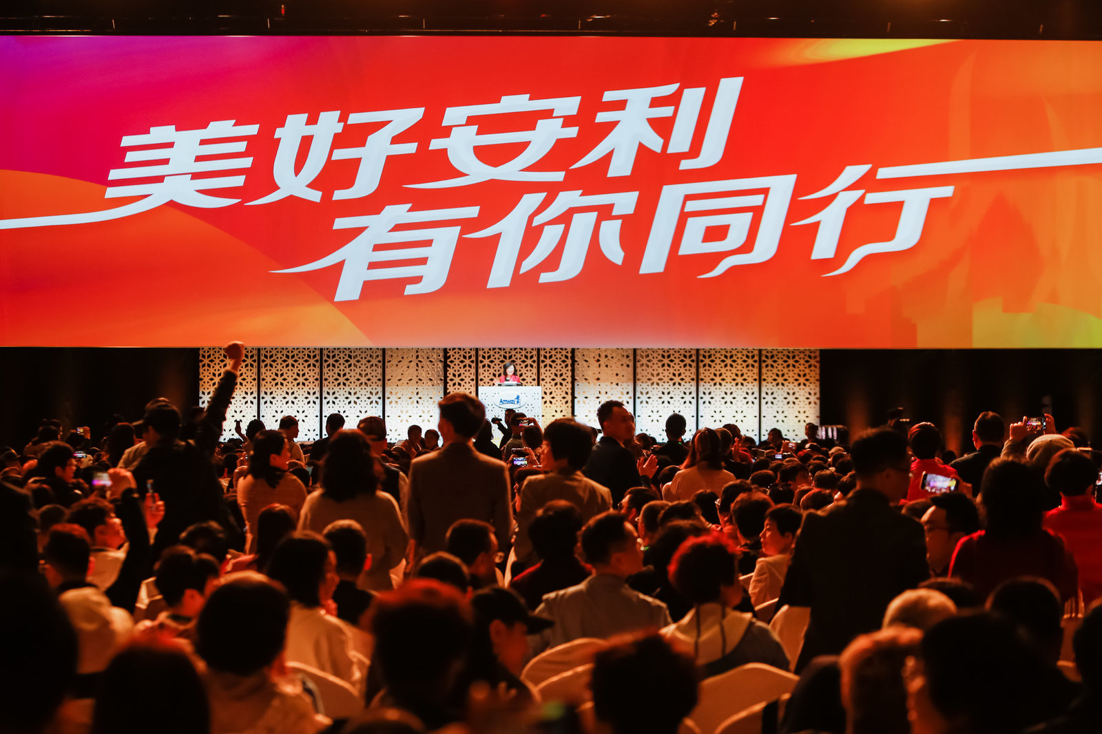活动现场巨幅大屏醒目展示“美好安利 有你同行”主题标语，台上设置安利品牌发言台，台下座无虚席，参会伙伴热情高涨，不少人举手机记录精彩内容，更有伙伴振奋举手响应，现场氛围热烈昂扬，尽显安利伙伴的凝聚力与共赴美好的正向风貌。
