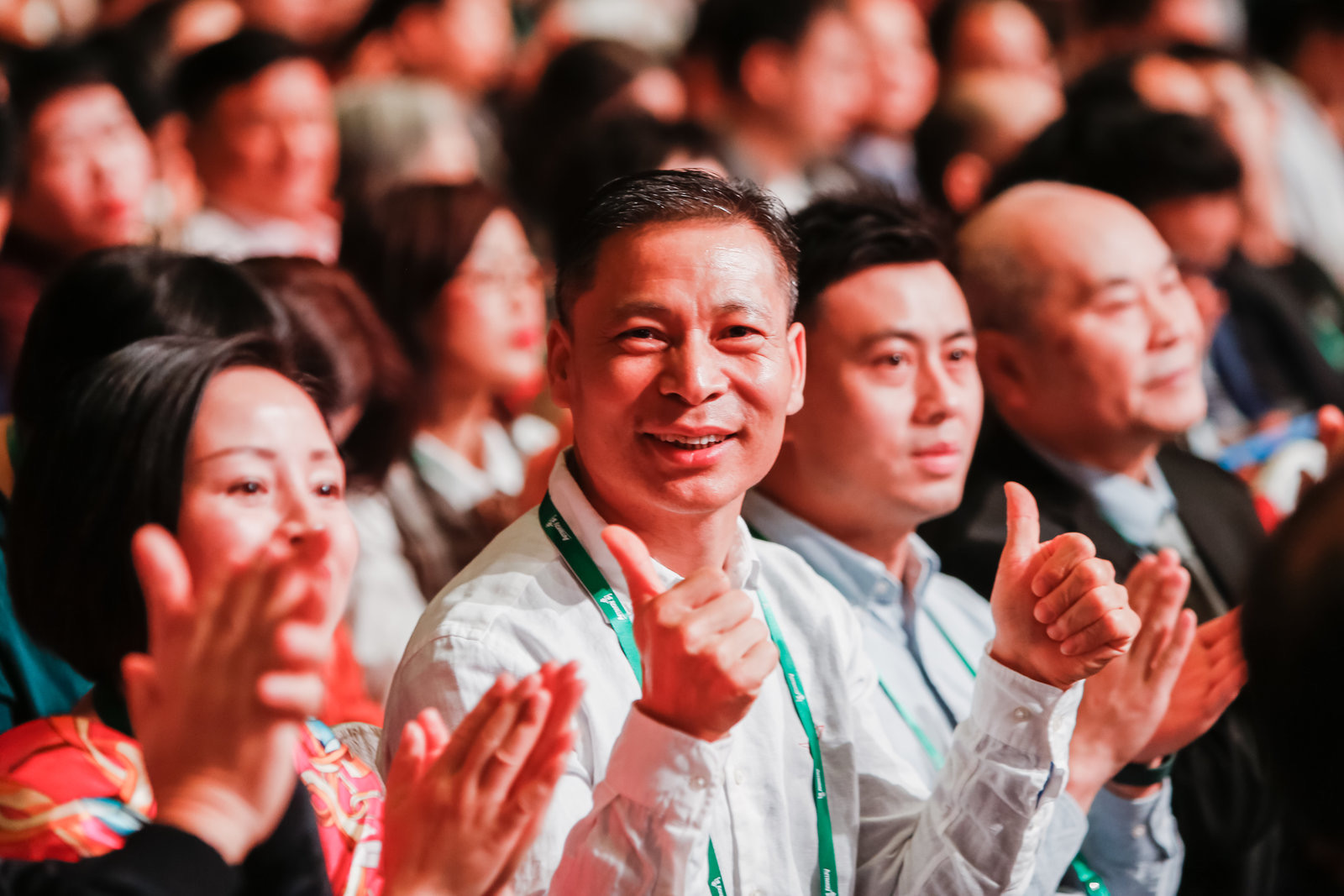 安利品牌活动现场，众多参会伙伴齐聚一堂，一位佩戴印有Amway标识绿色挂绳的男士面露喜色，竖起双手大拇指点赞，周围伙伴同步鼓掌致意，现场氛围热烈昂扬，充分展现出安利伙伴对活动的高度认可，以及团队积极向上的蓬勃风貌。