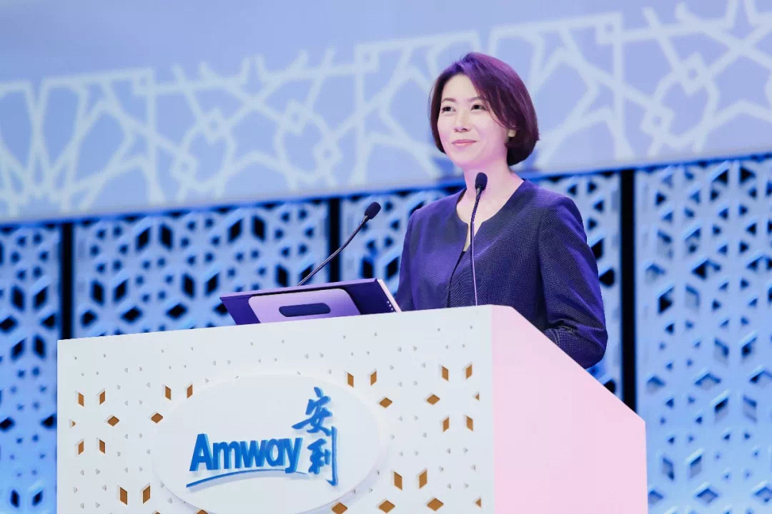 活动现场，身着正装的女性嘉宾站在印有“Amway 安利”标识的镂空设计演讲台后发言，演讲台配置双麦克风，背景为蓝调几何镂空造型的舞台布置，整体氛围专业庄重，彰显安利品牌活动的正式质感。