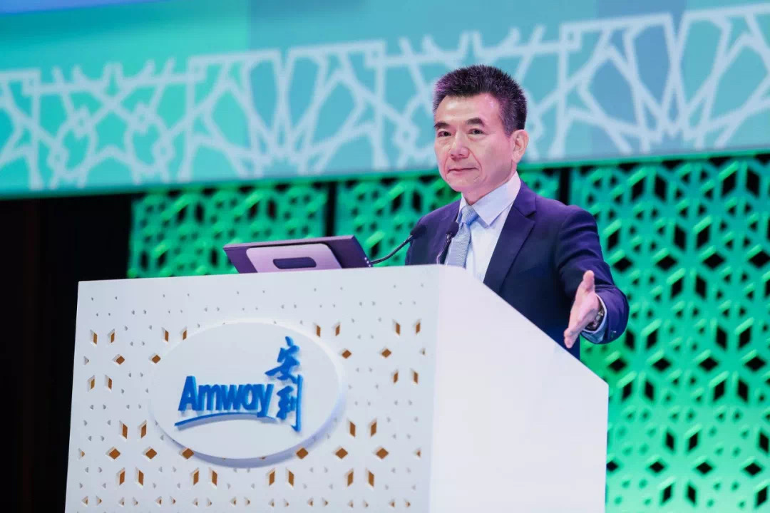 活动现场，身着正装的高管站在印有“Amway 安利”标识的镂空演讲台后发言，手部舒展做出手势，背景为青绿色几何纹样装饰大屏，整体氛围专业郑重，充分展现出安利积极正向的品牌风貌与活动的正式属性。