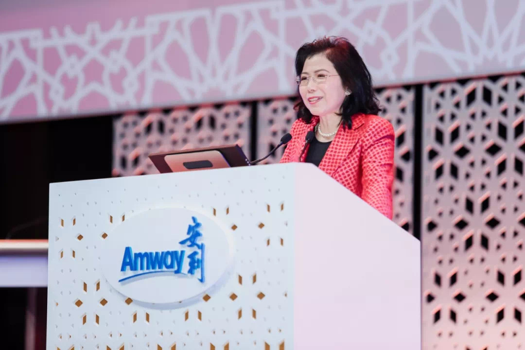 活动现场，身着红色格纹外套的女性高管站在印有“Amway 安利”标识的定制讲台后发表讲话，背景为设计感十足的镂空舞台，整体氛围专业庄重、积极明快，充分展现安利品牌活动的专业质感与正向风貌。
