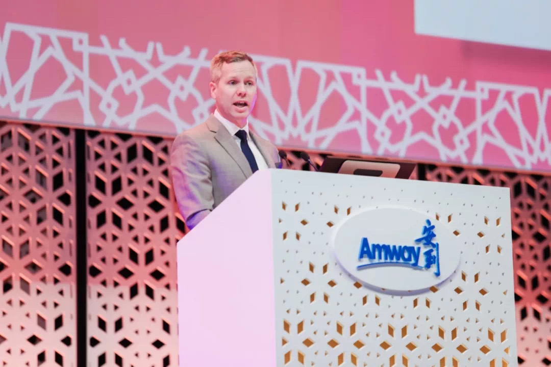 画面中身着正装的外籍高管正站在印有“Amway”“安利”标识的白色镂空演讲台后发言，背景是同系列几何纹样的粉调舞台布置，现场氛围正式专业，充分展现安利品牌活动的严谨风貌与正向氛围。