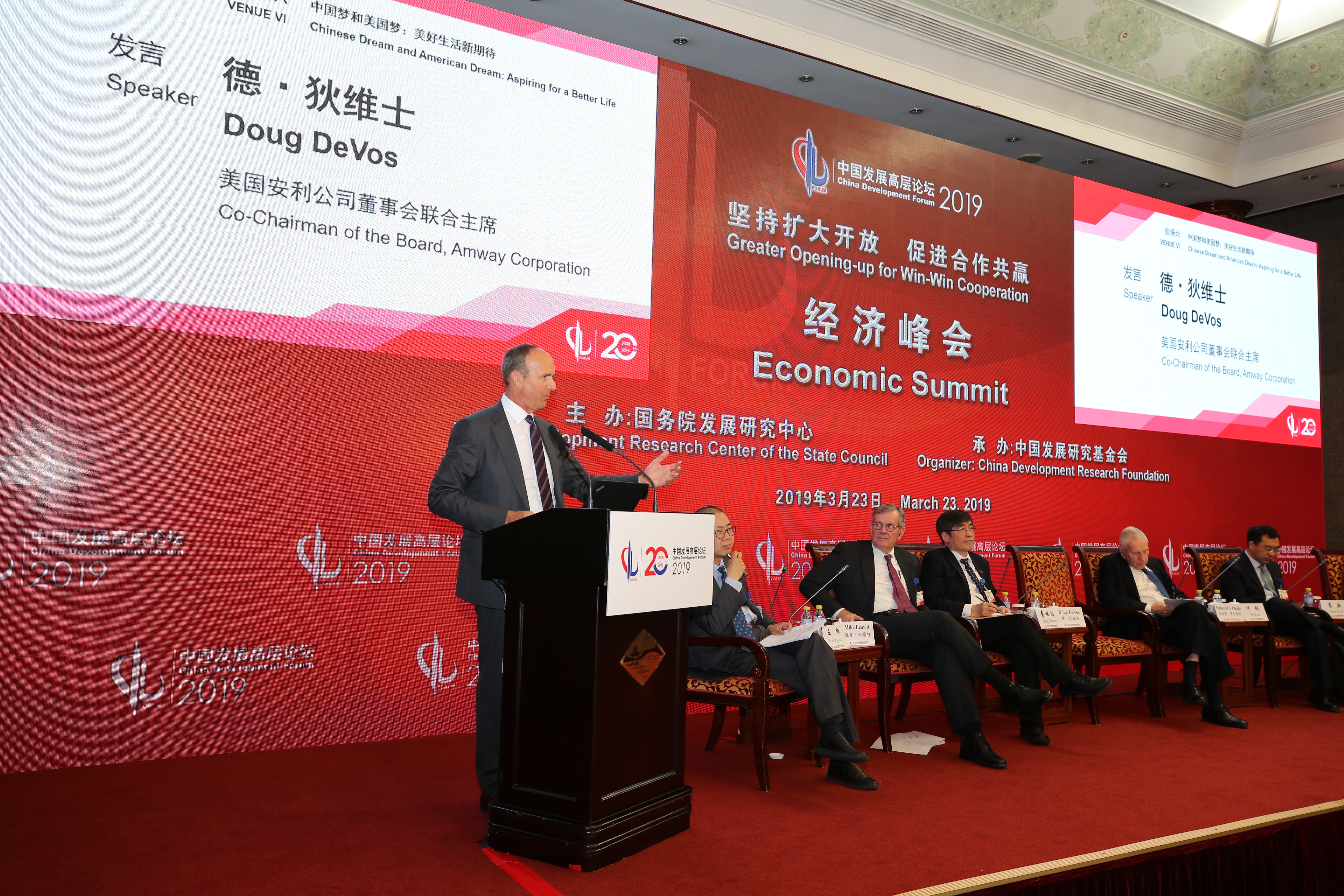本场景为2019中国发展高层论坛经济峰会现场，峰会主题为“坚持扩大开放 促进合作共赢”，美国安利公司董事会联合主席德·狄维士正在台上发言，台下就坐多位参会嘉宾，现场氛围庄重专业，彰显安利积极参与中外发展交流的态度。