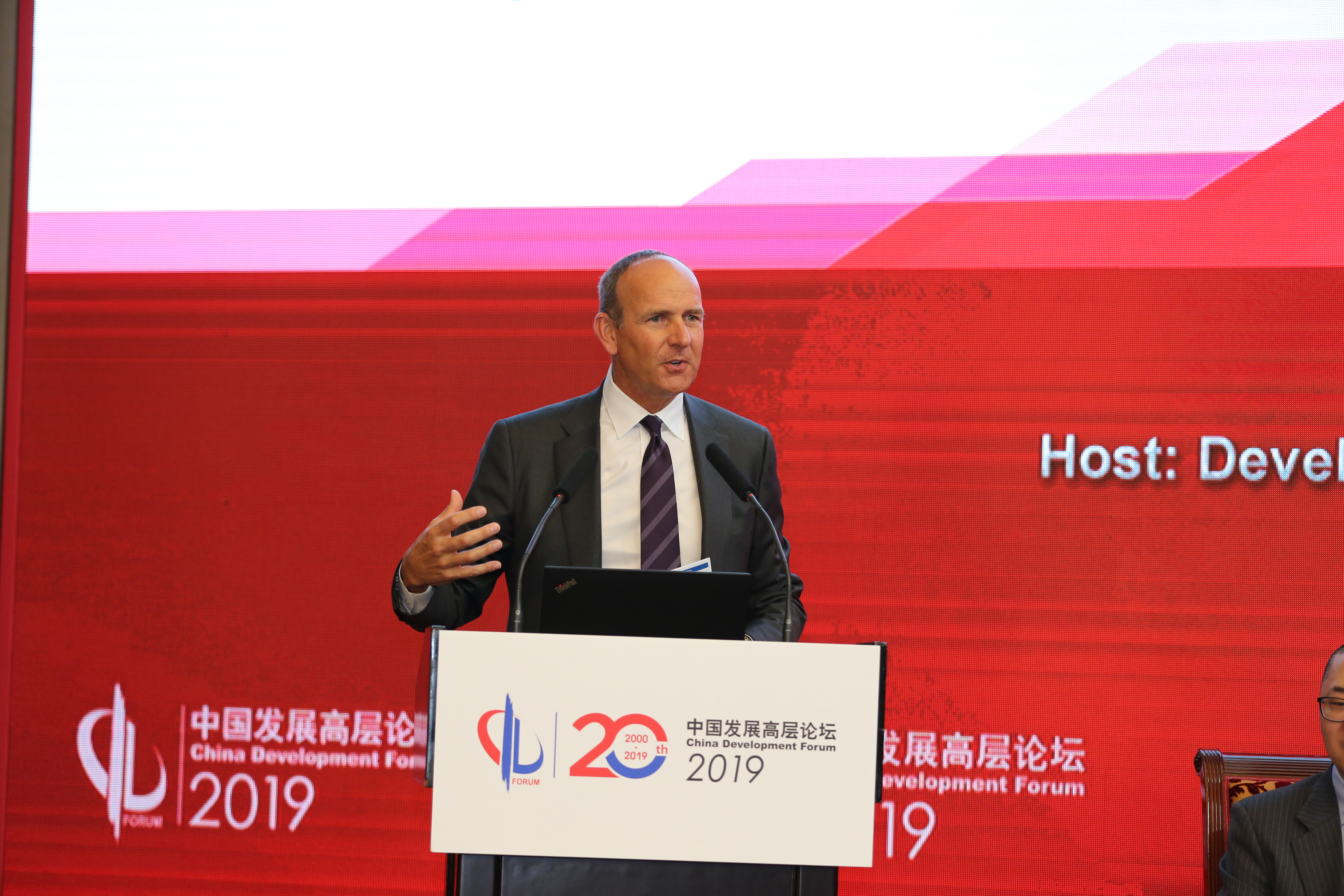 画面为2019中国发展高层论坛现场，演讲台标注有论坛2000-2019二十周年专属标识，外籍正装嘉宾正站在台前发言，手势舒展、语态从容，背景红色主题大屏庄重醒目，整体氛围专业正式，尽显高端论坛的权威属性。