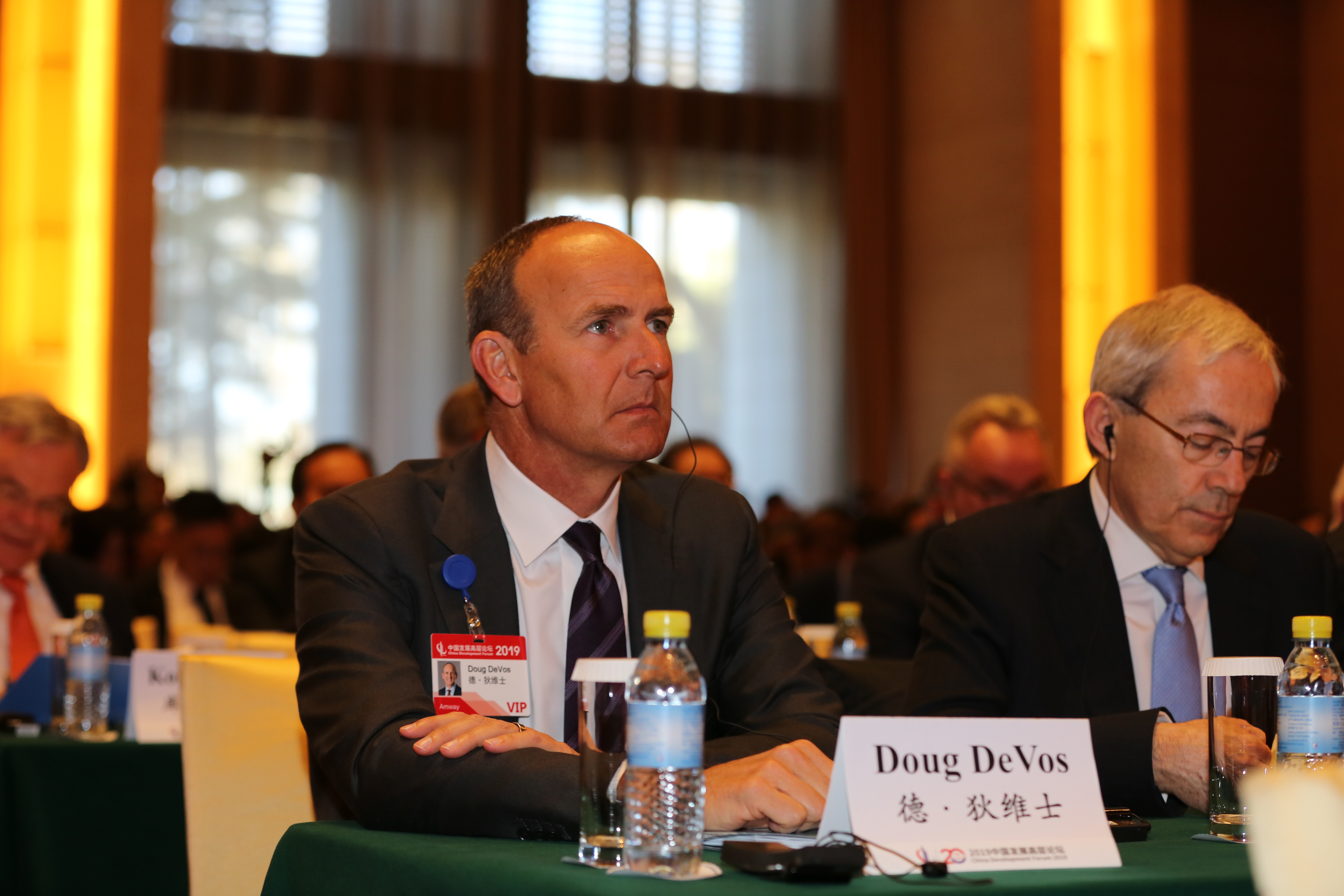 2019中国发展高层论坛现场，安利德·狄维士（Doug DeVos）佩戴VIP参会证件，身着正装端坐于会议桌前，神情专注认真，会场布置规整庄重，整体氛围专业肃穆，展现出企业高层对行业交流研讨的重视。