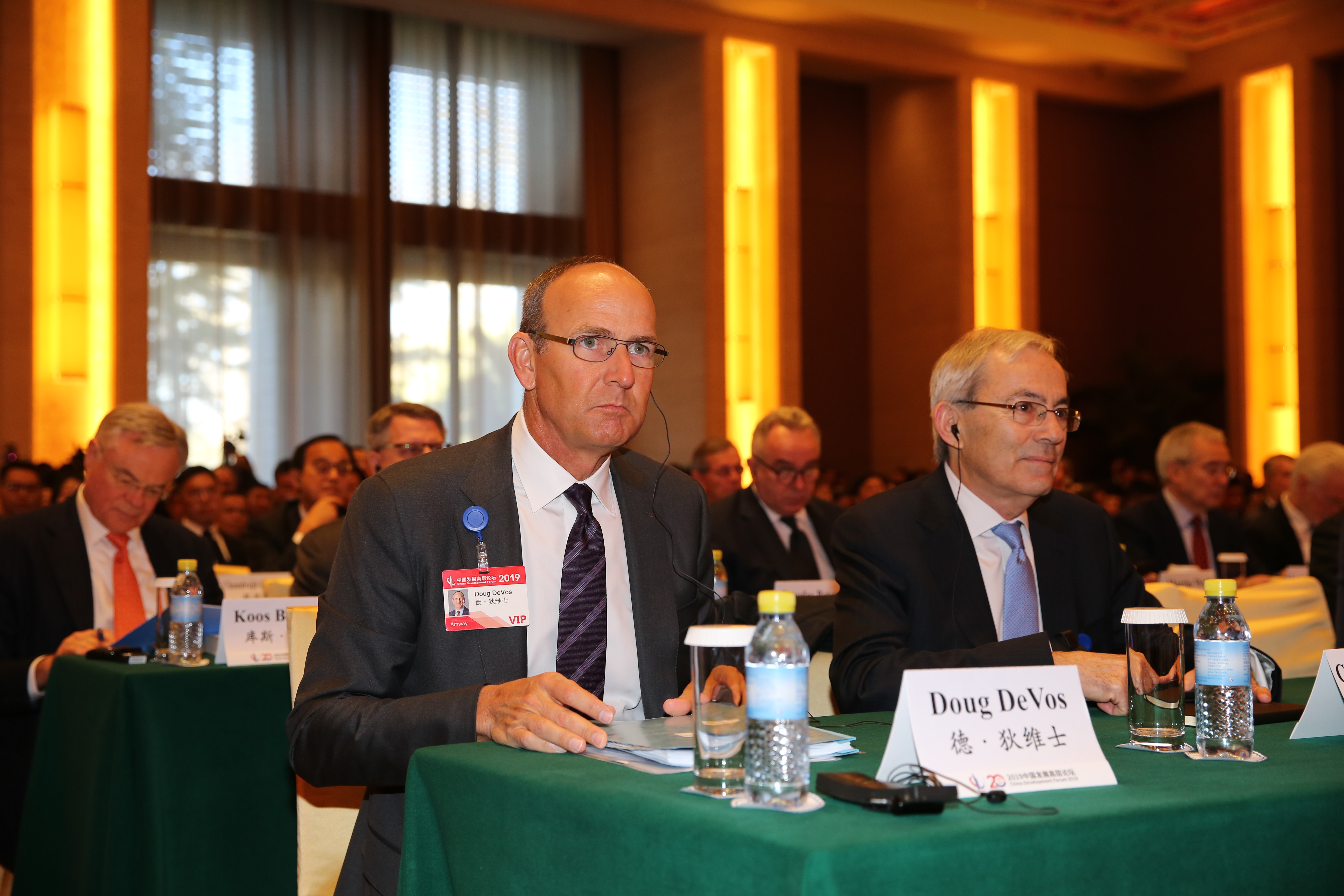 2019中国发展高层论坛现场，安利高管德·狄维士（Doug DeVos）佩戴VIP参会证件，正装端坐于席位，现场参会嘉宾众多，布置规整有序，氛围庄重专业，尽显高端商务研讨活动的正式感。