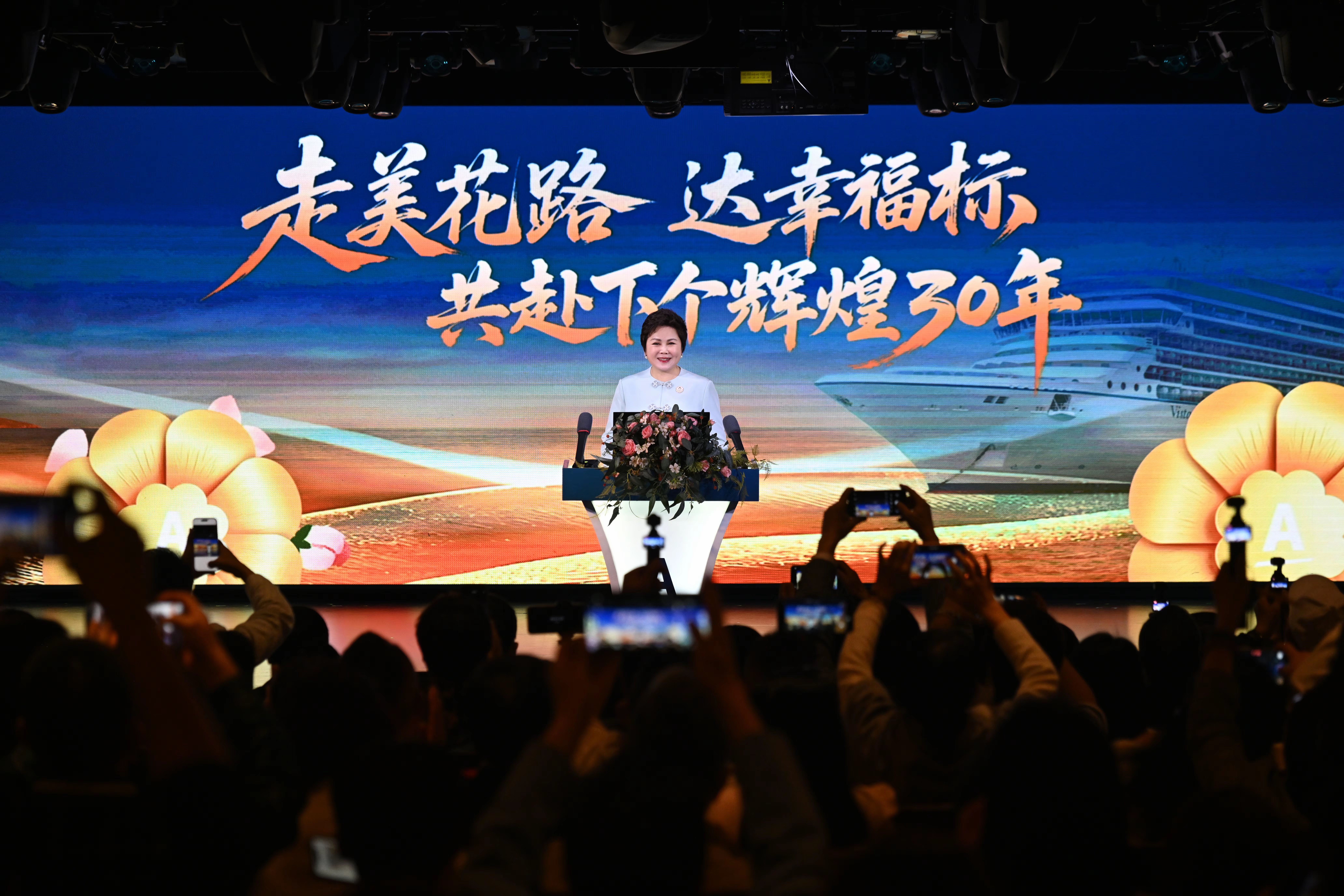 活动现场背景大屏显示“走美花路 达幸福标 共赴下个辉煌30年”主题字样，搭配游轮、花朵视觉元素，领导在台前致辞，台下参会伙伴纷纷举起手机记录精彩时刻，现场氛围隆重热烈，尽显安利积极向上的发展风貌。