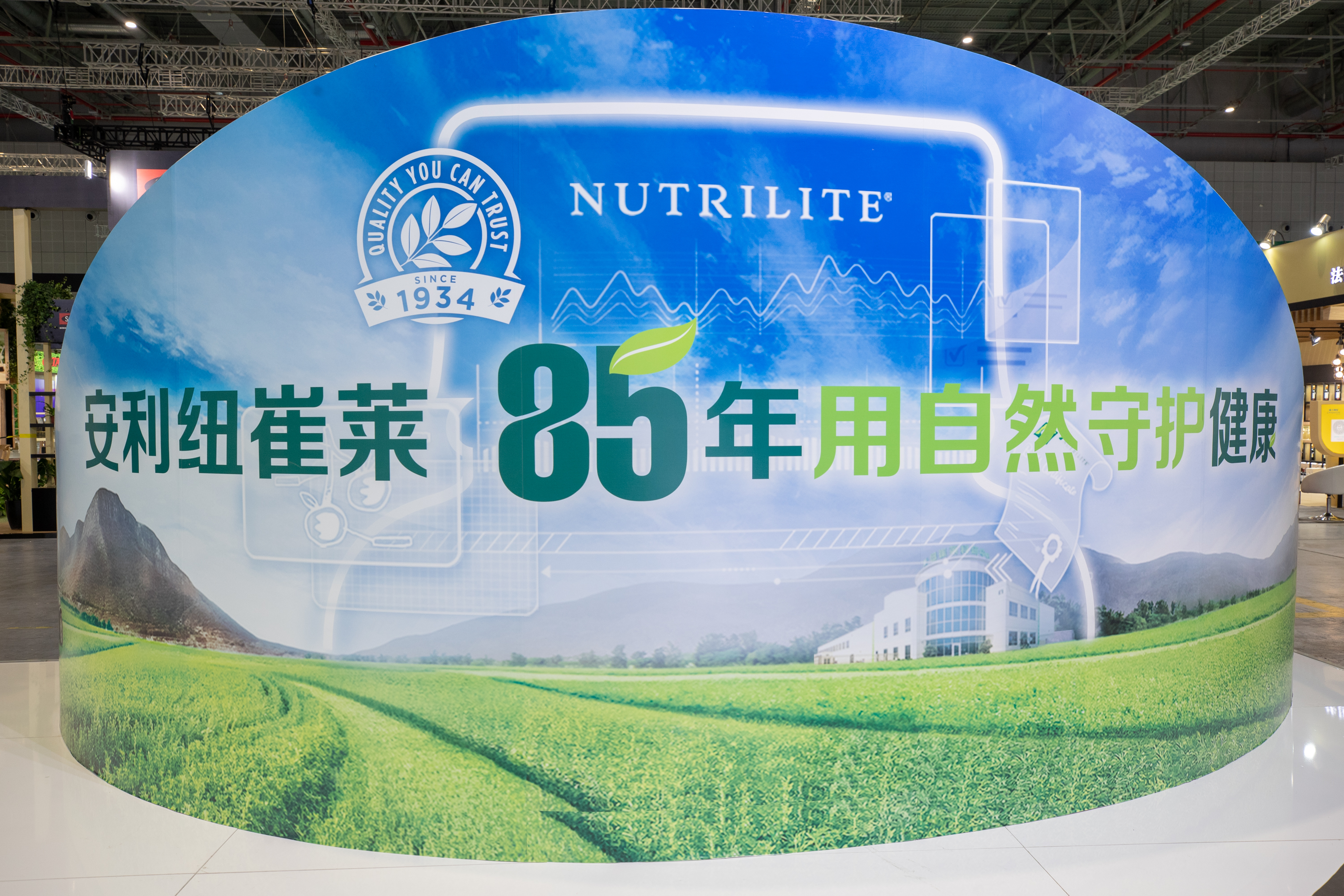 这是展会现场的安利纽崔莱弧形宣传背景板，板面上印有“NUTRILITE”“QUALITY YOU CAN TRUST SINCE 1934”品牌标识，以及核心标语“安利纽崔莱85年用自然守护健康”，搭配有机农场、蓝天远山的清新画面，风格明快积极，传递品牌自然健康的理念。