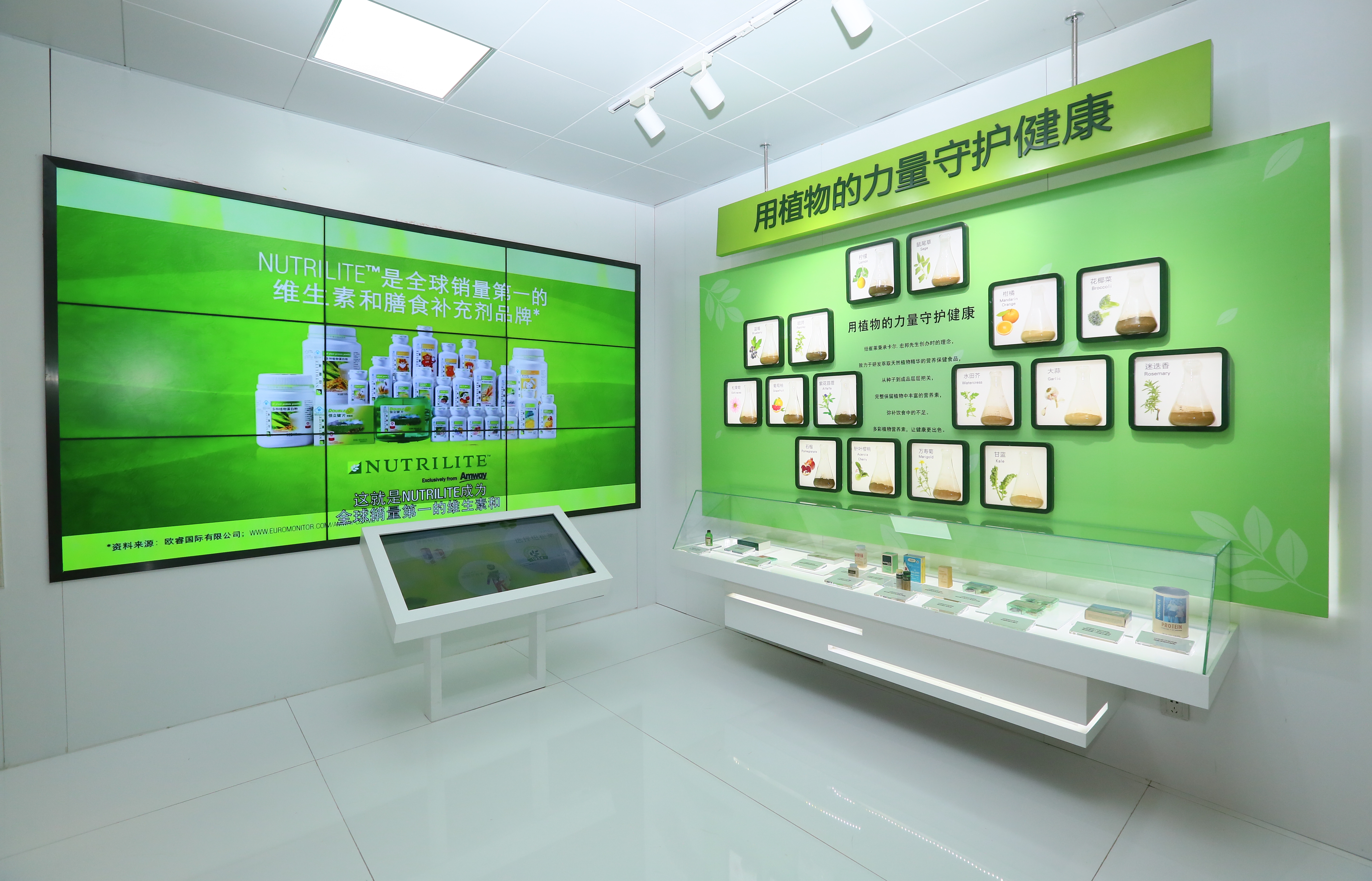 展厅环境整洁明亮，墙面大屏标注“NUTRILITE是全球销量第一的维生素和膳食补充剂品牌”，展示纽崔莱全系列营养产品；另一侧绿色展墙标注“用植物的力量守护健康”，陈列植物原料科普内容，下方玻璃展柜摆放相关产品，搭配智能查询设备，尽显品牌专业健康的形象。