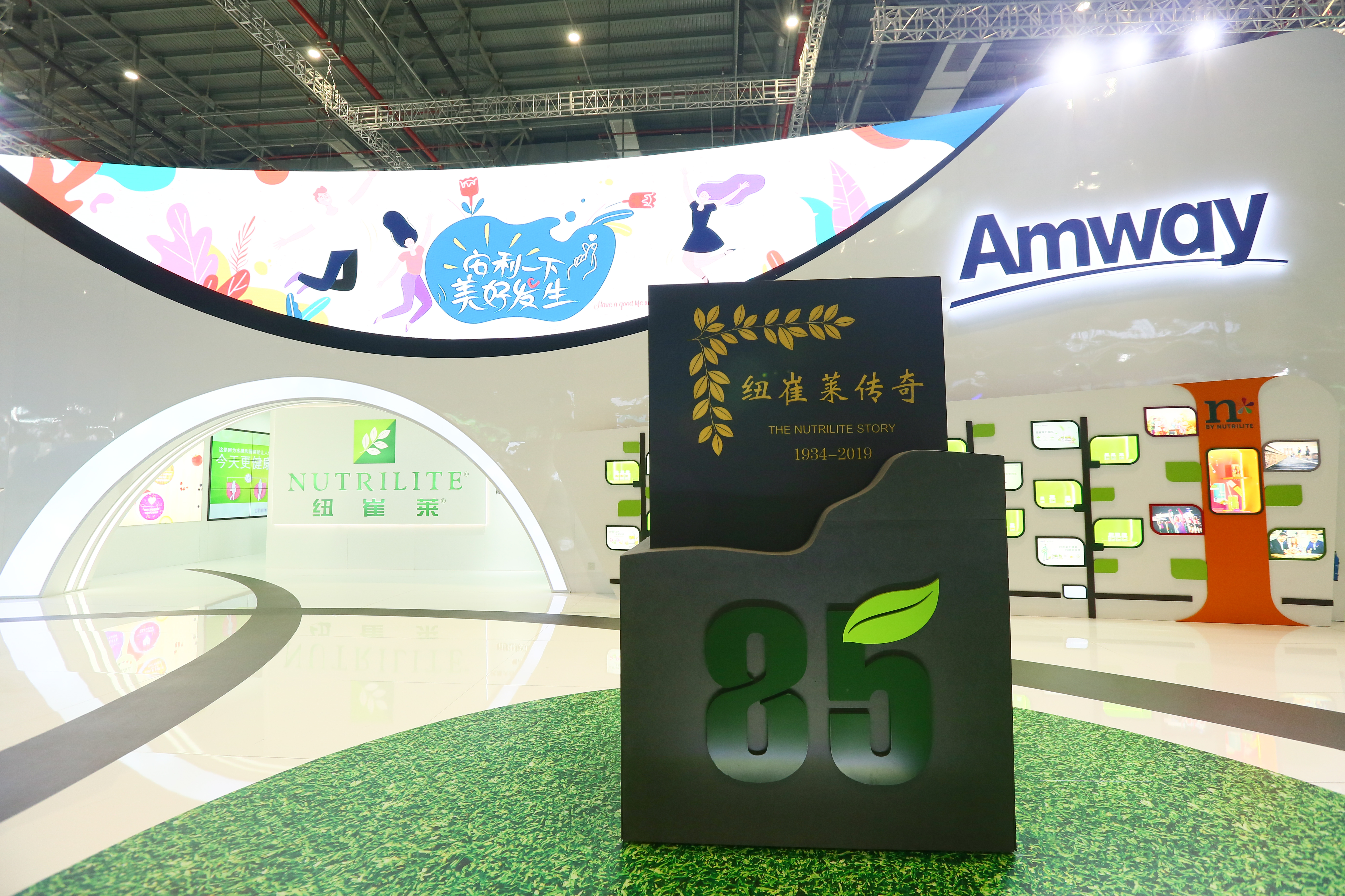 画面为安利活动现场展示区，前景立有纽崔莱85周年纪念装置，上方标识牌标注“纽崔莱传奇 THE NUTRILITE STORY 1934-2019”。背景弧形大屏印有“安利一下 美好发生”标语，墙面设有Amway、纽崔莱品牌标识与内容展示区，整体场地明亮清新，尽显品牌健康活力的积极调性。