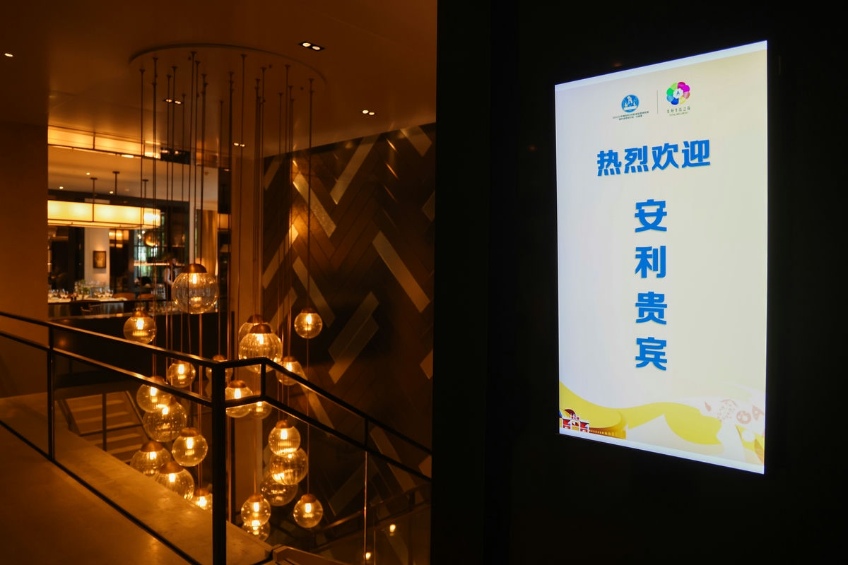 画面右侧电子屏清晰显示“热烈欢迎 安利贵宾”字样，旁侧活动场地装修精致考究，暖光玻璃球形吊灯错落垂落，整体氛围雅致庄重、热忱贴心，充分彰显对安利参会贵宾的礼遇，契合品牌活动的高端质感。