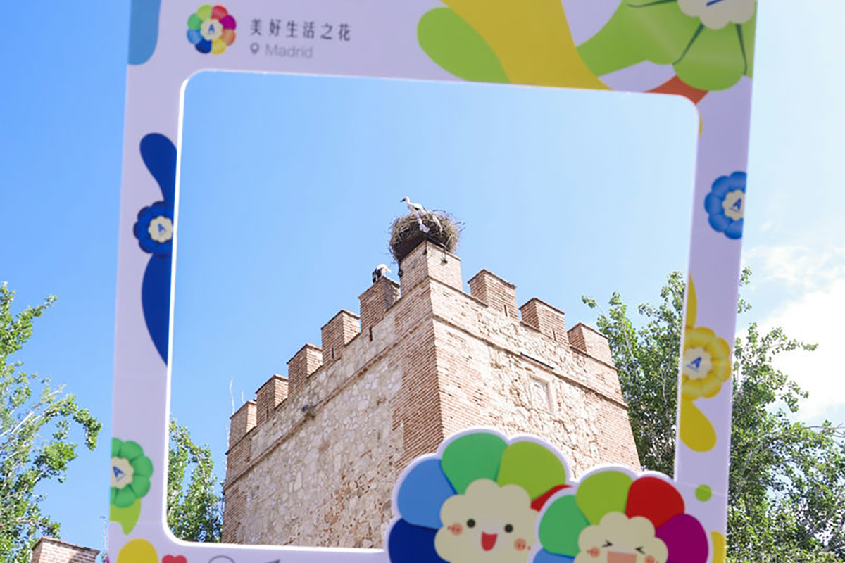 印有“美好生活之花”“Madrid”字样的安利定制打卡框置于户外，框内呈现马德里砖石古堡景致，古堡顶端有鹳鸟栖息在巢穴中，周边是澄澈蓝天与青葱绿树，氛围明快惬意，传递出美好鲜活的活动质感。