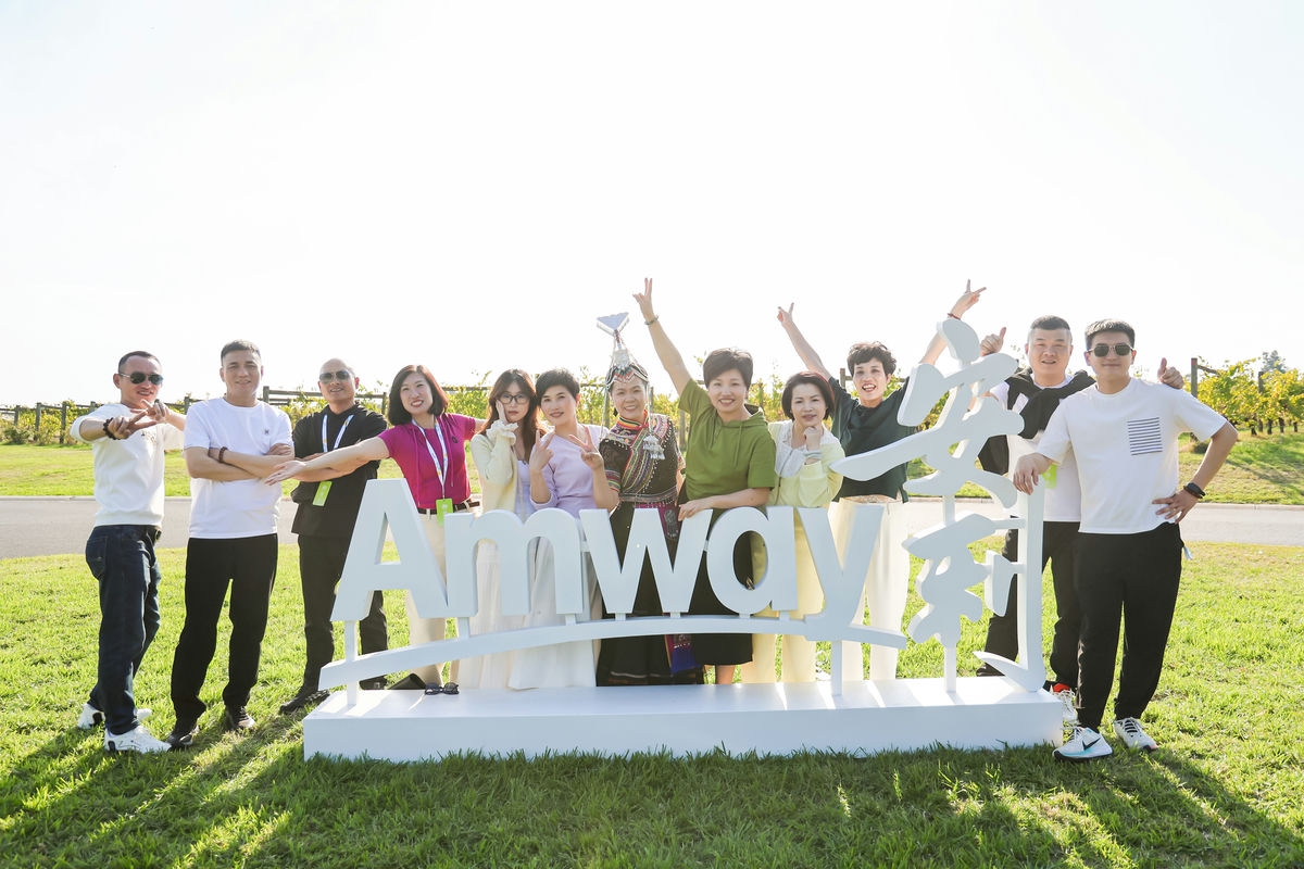 晴朗户外的青草地中，十余位安利伙伴站在印有“Amway”“安利”字样的立体品牌标识旁合影，大家神态轻松愉悦、笑容灿烂，还有身着民族服饰的伙伴参与其中，整体氛围活泼温暖，尽显安利伙伴的凝聚力与积极向上的品牌风貌。