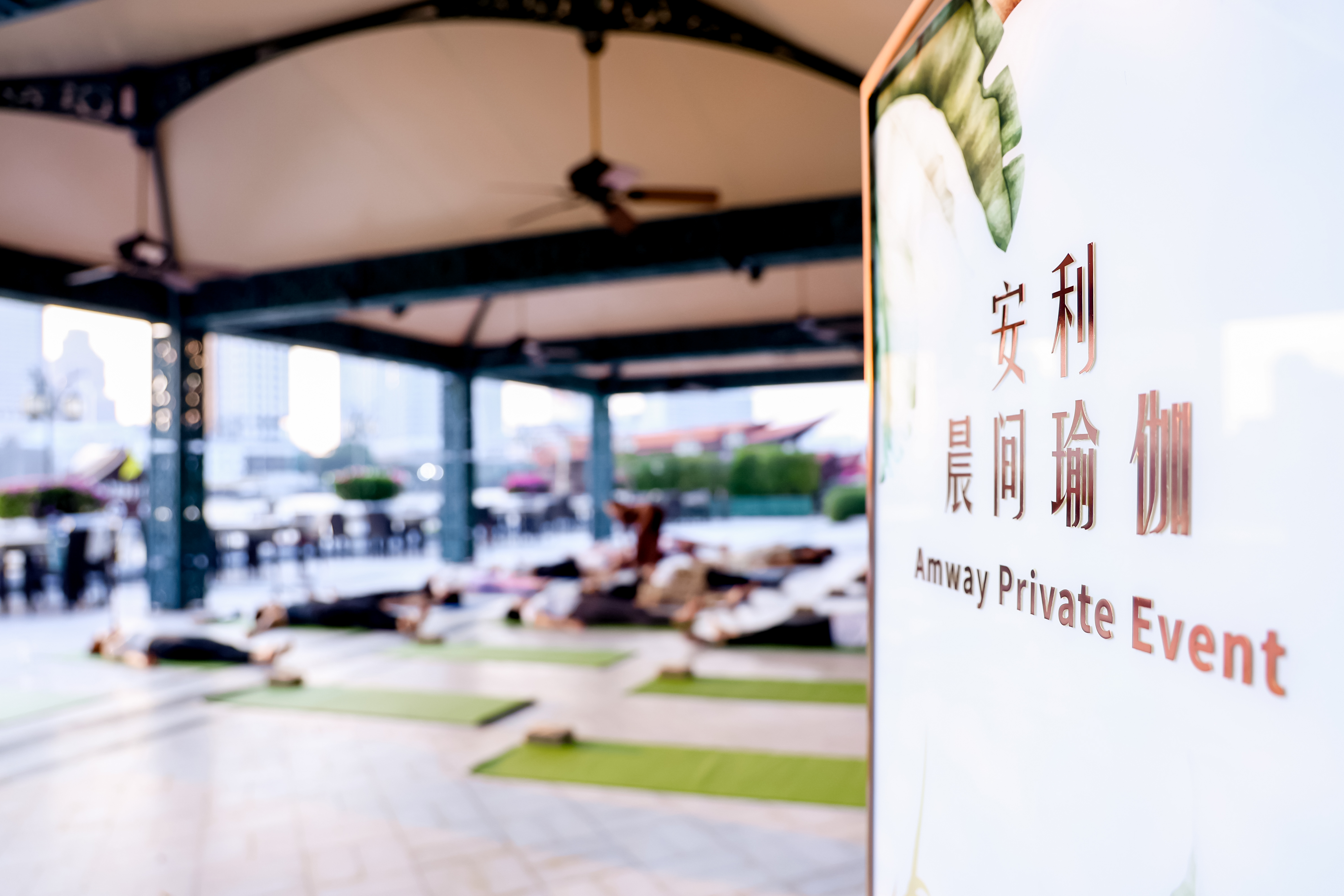 画面右侧立牌印有“安利 晨间瑜伽”“Amway Private Event”字样，背景开阔的户外场地中整齐排布着瑜伽垫，参与活动的伙伴们正舒展身姿练习瑜伽，氛围舒缓放松，尽显安利倡导健康美好生活的品牌理念。