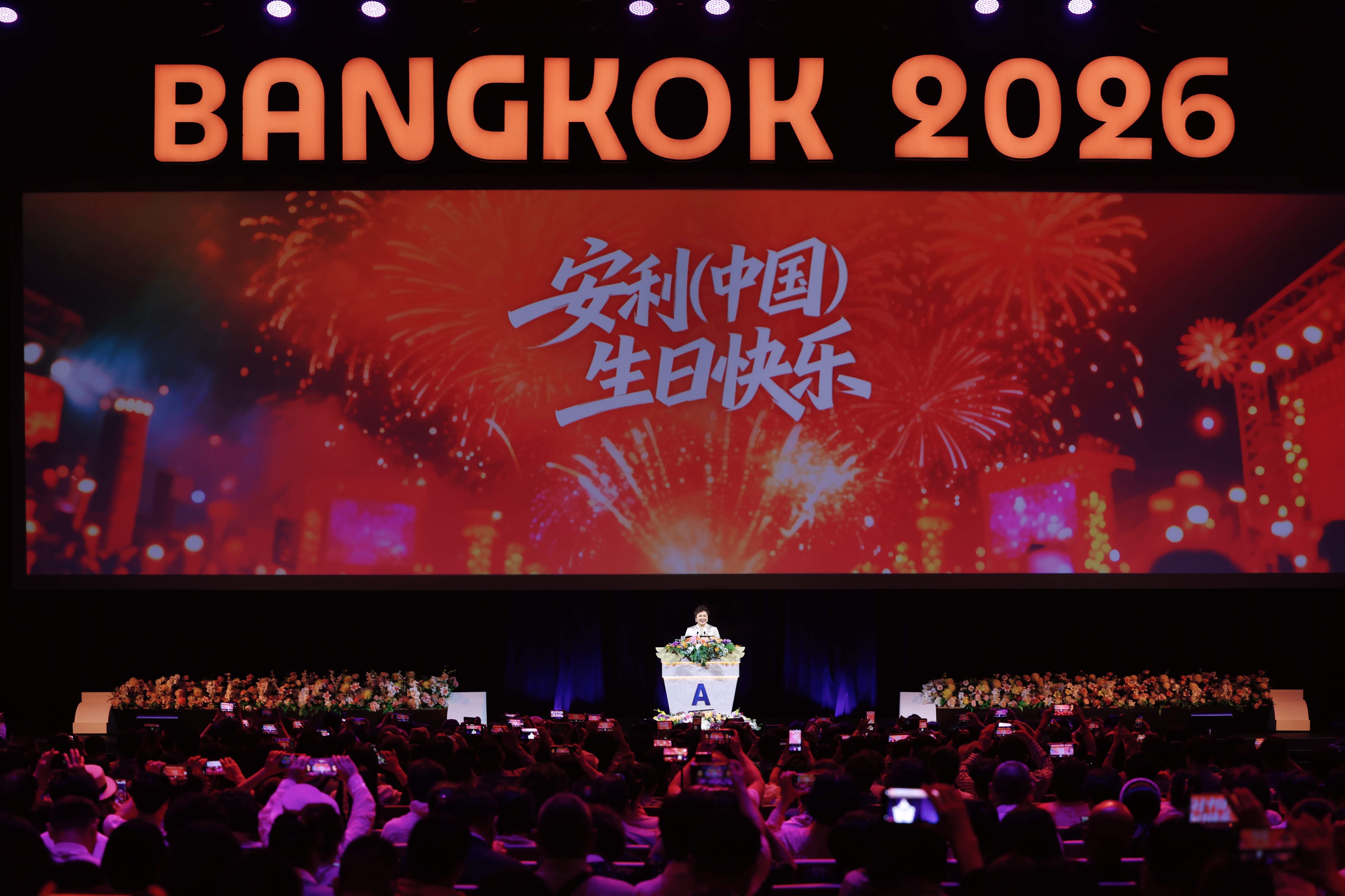 活动现场上方醒目标注“BANGKOK 2026”字样，主屏幕以绚烂烟花为背景，呈现“安利(中国)生日快乐”的暖心祝福，发言人在印有品牌标识的讲台致辞，台下参会伙伴纷纷举起手机记录精彩时刻，全场氛围热烈喜庆，尽显安利大家庭的温暖凝聚力。