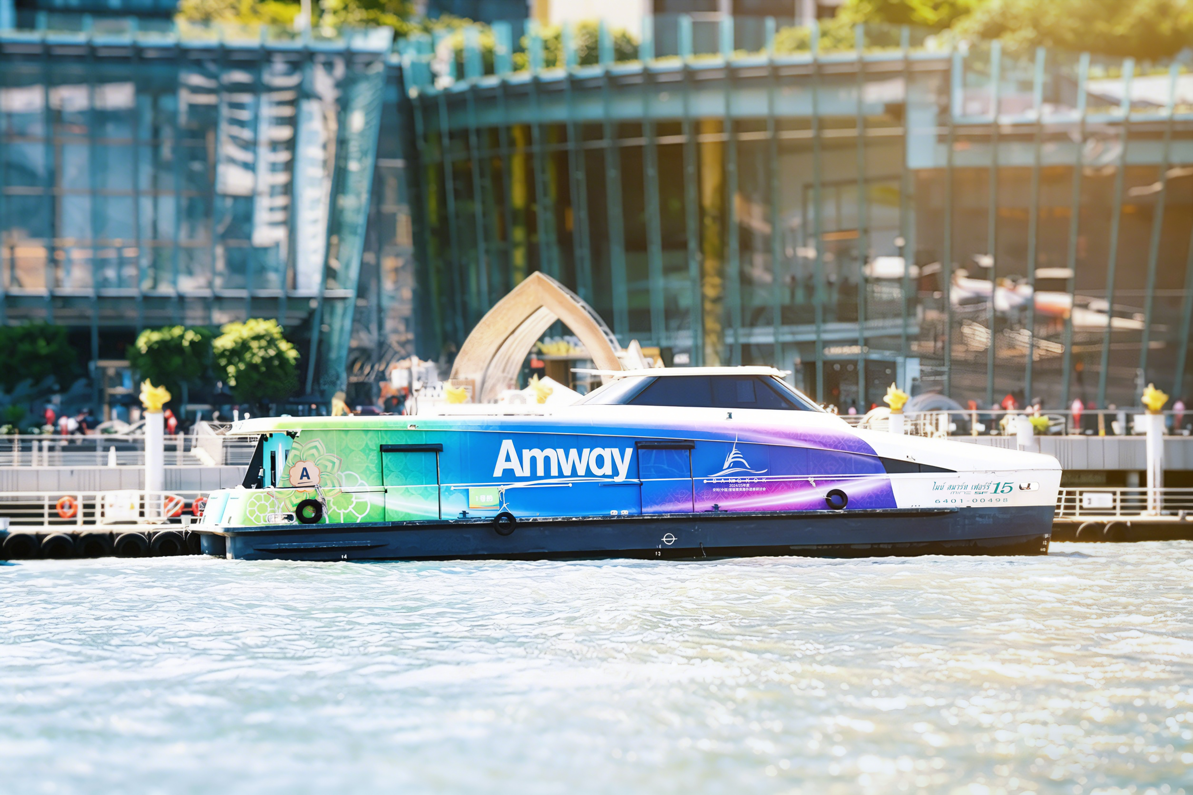 画面中印有醒目「Amway」标识的定制游船，采用绿蓝紫渐变特色涂装，停靠在波光粼粼的河畔码头，背景是时尚现代的城市滨水建筑，整体氛围明快鲜亮，作为安利曼谷品牌活动的专属水上交通工具，尽显品牌活力与专属礼遇质感。