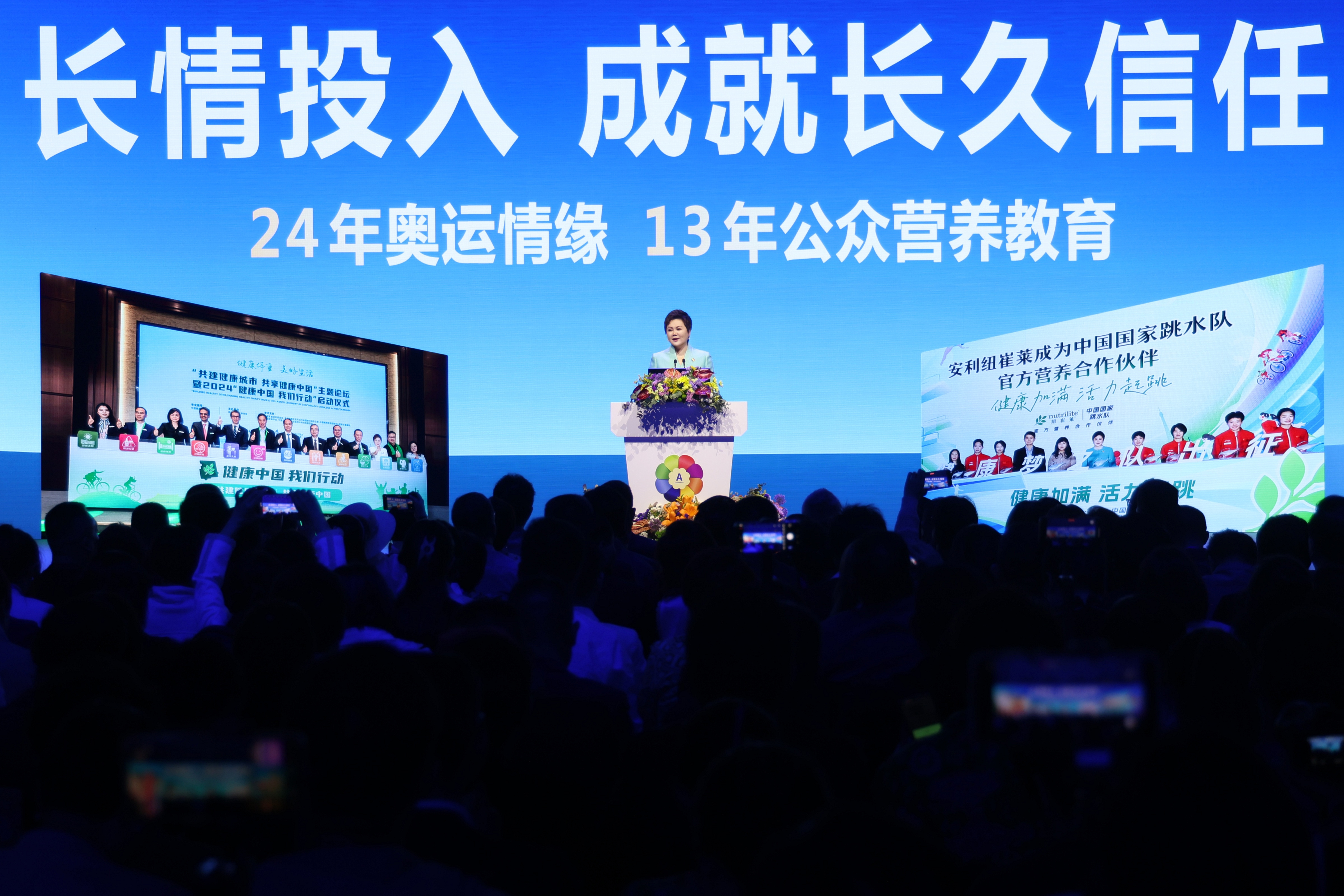 活动主背景标注“长情投入 成就长久信任”“24年奥运情缘 13年公众营养教育”，本次为2024“健康中国 我们行动”启动仪式现场，侧屏展示安利纽崔莱为中国国家跳水队官方营养合作伙伴，发言人在台上致辞，台下参会人员齐聚，氛围庄重热烈，尽显安利深耕健康领域的品牌担当。