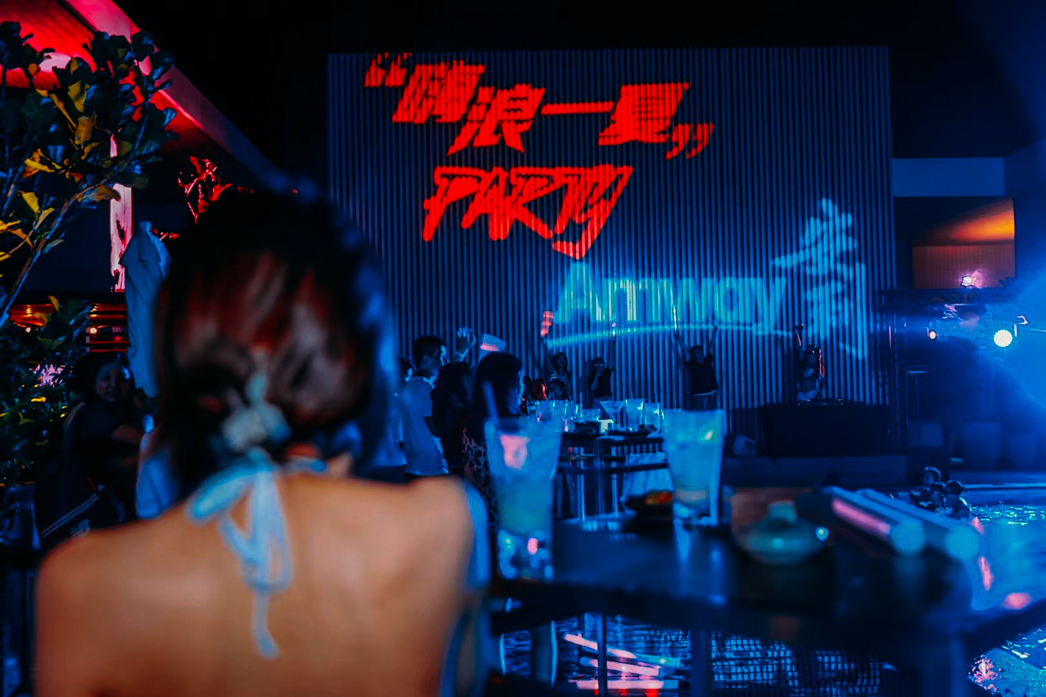 现场大屏醒目展示“嗨浪一夏”“PARTY”与“Amway”字样，蓝红交织的霓虹灯光氛围感拉满，场内摆放饮品的桌台错落分布，参与活动的伙伴齐聚现场，氛围热烈欢快，尽显安利年轻活力的品牌调性。