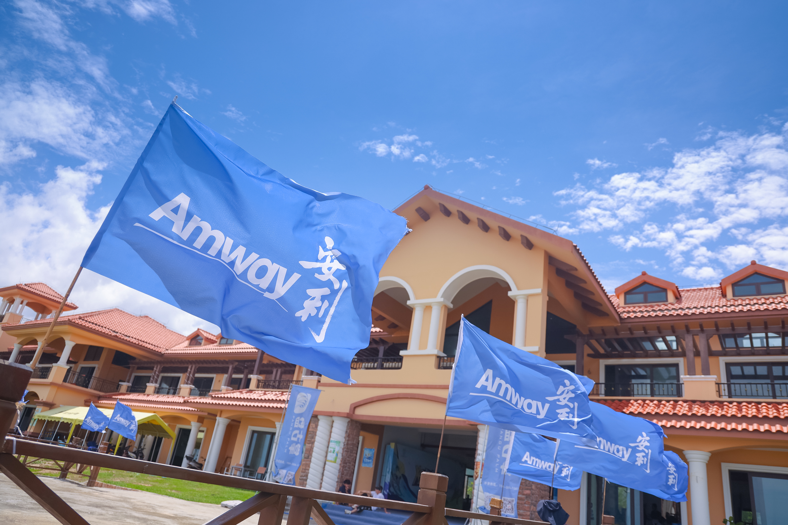 澄澈蓝天白云下，暖黄色红顶的度假风建筑前，多面印有“Amway”“安利”字样的蓝色品牌旗帜迎风舒展，整体场景明亮开阔、朝气蓬勃，尽显安利品牌活动的活力与积极向上的氛围。