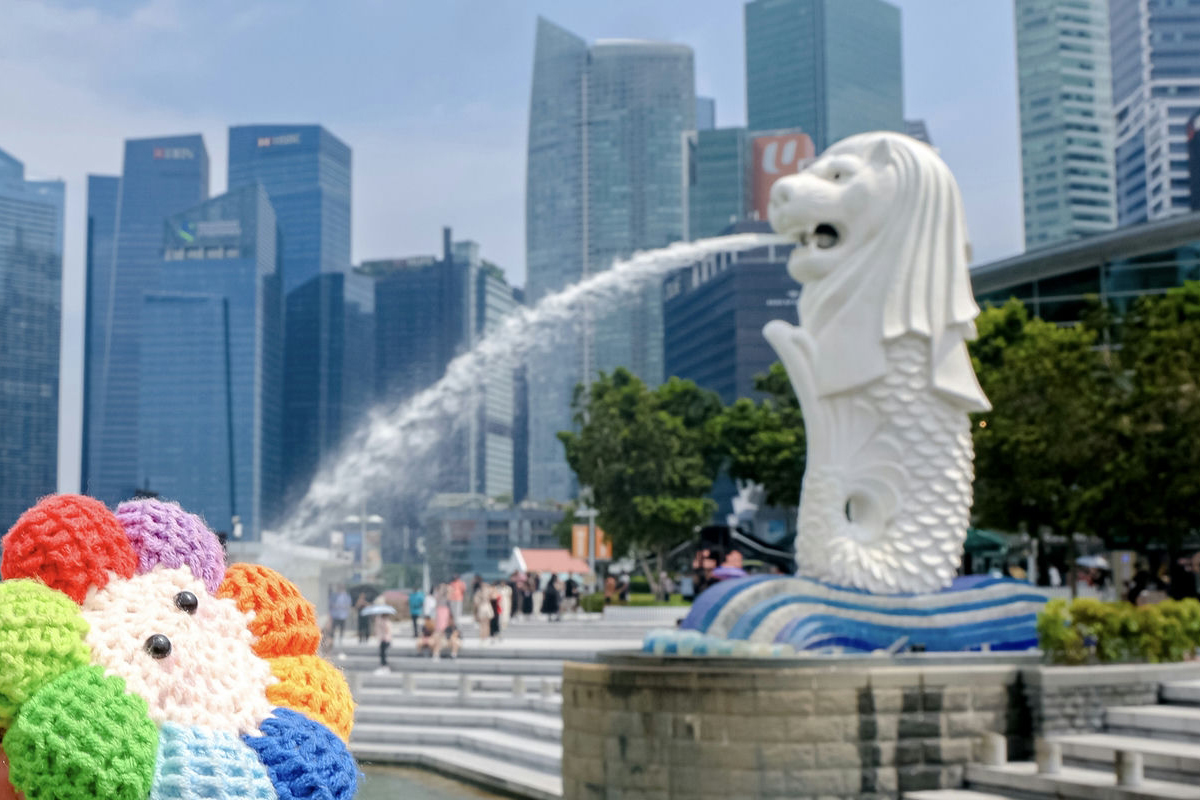 画面前景是色彩缤纷的针织太阳花手作，背景为新加坡标志性鱼尾狮喷泉，滨海湾摩天楼群矗立后方，广场上游人往来，晴日下氛围明快鲜活，尽显安利海外行程的精彩活力与满满朝气。