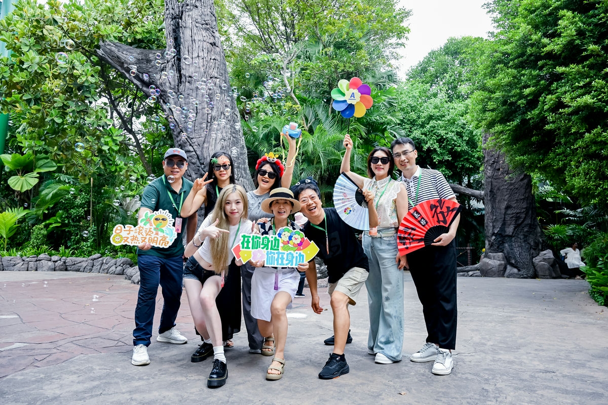 安利户外团建活动现场，葱郁绿植环绕，多位伙伴笑容灿烂齐聚合影。大家手持“日子滚烫 又热又明亮”“在你身边 你在身边”趣味手牌，搭配风车、折扇、泡泡机等道具，氛围轻松愉悦，尽显伙伴们昂扬鲜活的状态与融洽的团队氛围。