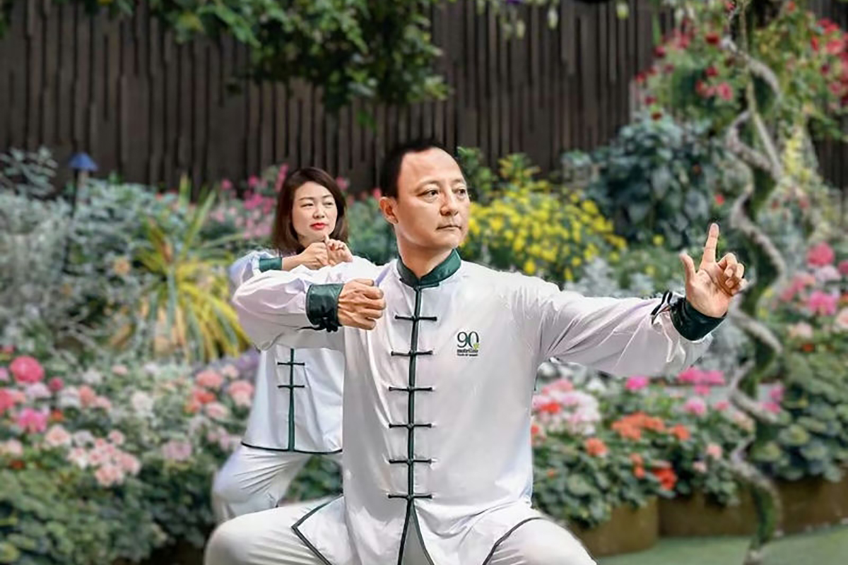画面中两位身着印有“90”标识中式太极服的安利活动参与者，在花木繁茂的户外场地扎马步习练太极，动作舒展、神态专注，氛围活力闲适，充分展现了安利倡导的健康积极的生活理念。