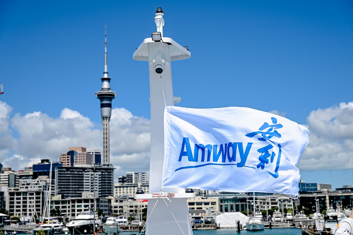 画面中印有“Amway 安利”字样的白色旗帜在游艇桅杆上迎风舒展，背景是奥克兰标志性天空塔，澄澈蓝天之下，滨海城市建筑错落、港口游艇排布，整体氛围开阔明朗，尽显安利海外活动的昂扬活力与开阔格局。