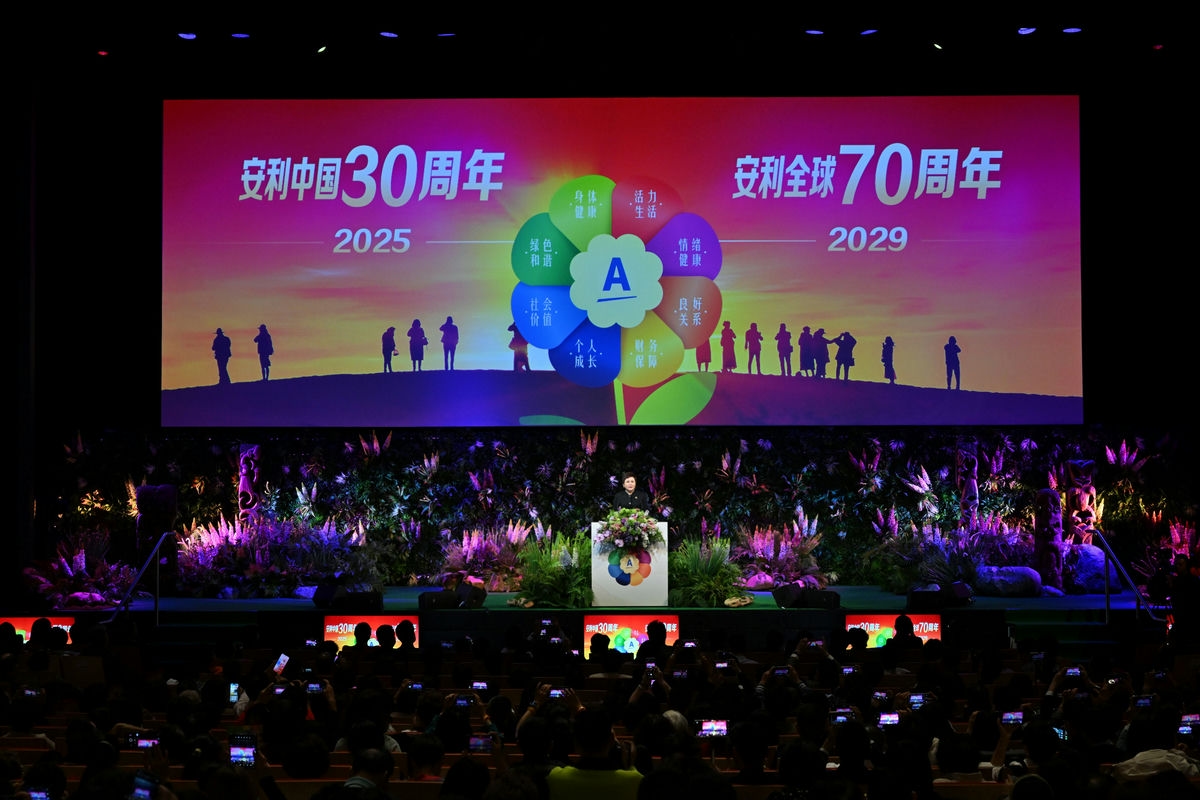 活动现场主屏幕展示“安利中国30周年 2025”“安利全球70周年 2029”字样，以及涵盖身体健康、活力生活等维度的品牌理念标识，演讲人在花艺装饰的台前宣讲，台下参会者纷纷举手机记录，现场氛围热烈庄重，彰显安利品牌正向发展风貌。