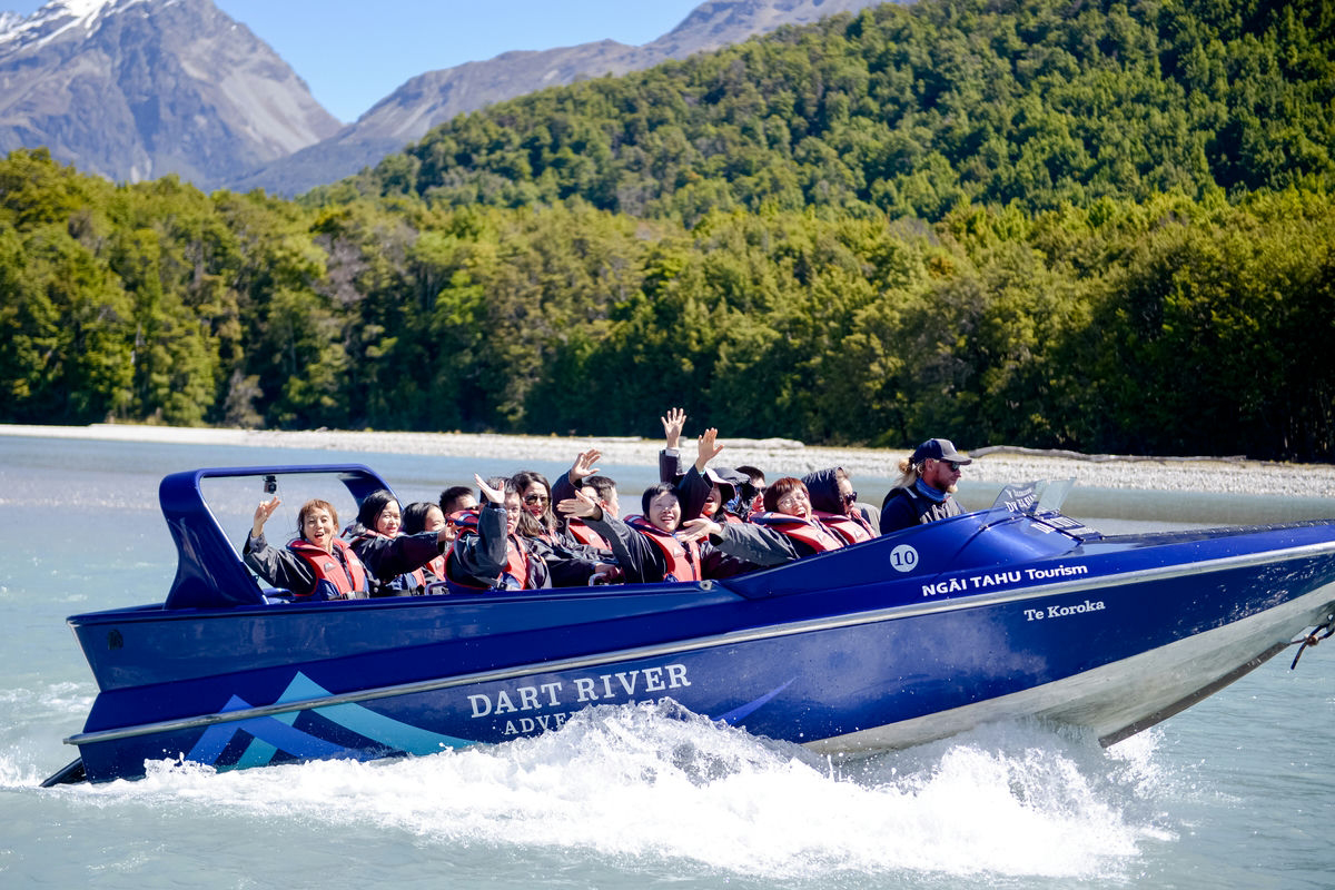 安利伙伴们身着红色救生衣，乘坐印有“DART RIVER”“NGĀI TAHU Tourism”“Te Koroka”字样的蓝色快艇在河面疾驰，大家笑容灿烂纷纷挥手，背景是覆雪山峦与葱郁林野，氛围明快热烈，尽显安利伙伴的昂扬活力与活动的愉悦质感。