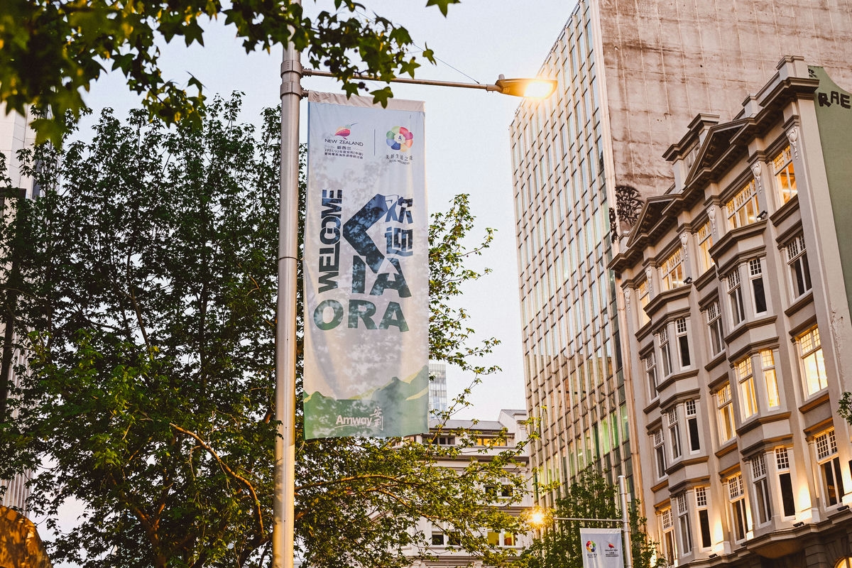 画面展示城市街道路灯杆上悬挂的安利活动主题道旗，道旗印有“WELCOME KIA ORA”“欢迎”“Amway安利”“NEW ZEALAND”“美好生活之夜”字样，旁侧绿植葱郁，周边错落着现代与复古风格建筑，氛围温暖热忱，尽显对活动参与者的欢迎心意。