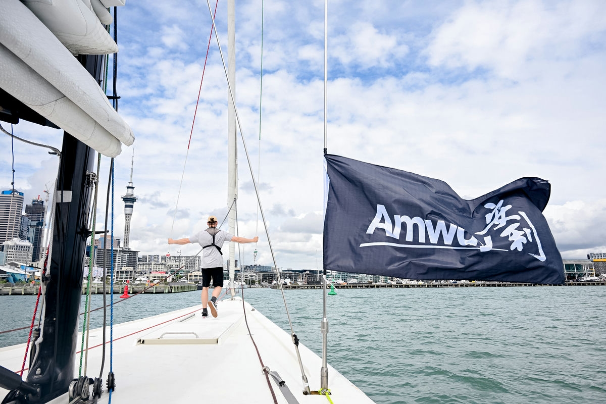 本次安利海上主题活动中，帆船航行于开阔海面，船侧悬挂印有“Amway”“澎湃”字样的品牌旗帜，一名伙伴立于船头舒展双臂，背景是奥克兰城市天际线与标志性天空塔，天朗气清海波荡漾，氛围昂扬向上，尽显安利伙伴积极探索的奋进风貌。